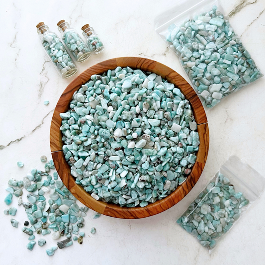 A wooden bowl filled with light blue and white Larimar Crystal Chips is surrounded by similar stones in plastic bags and small glass bottles. Additional stones, perfect for crafting supplies or metaphysical uses, are scattered on the white marble surface around the bowl.