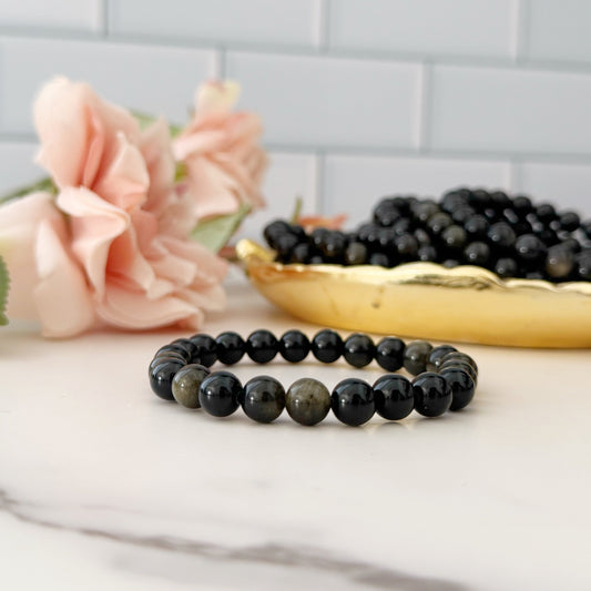 Black beaded bracelet on a marble surface with flowers and a gold dish in the background.