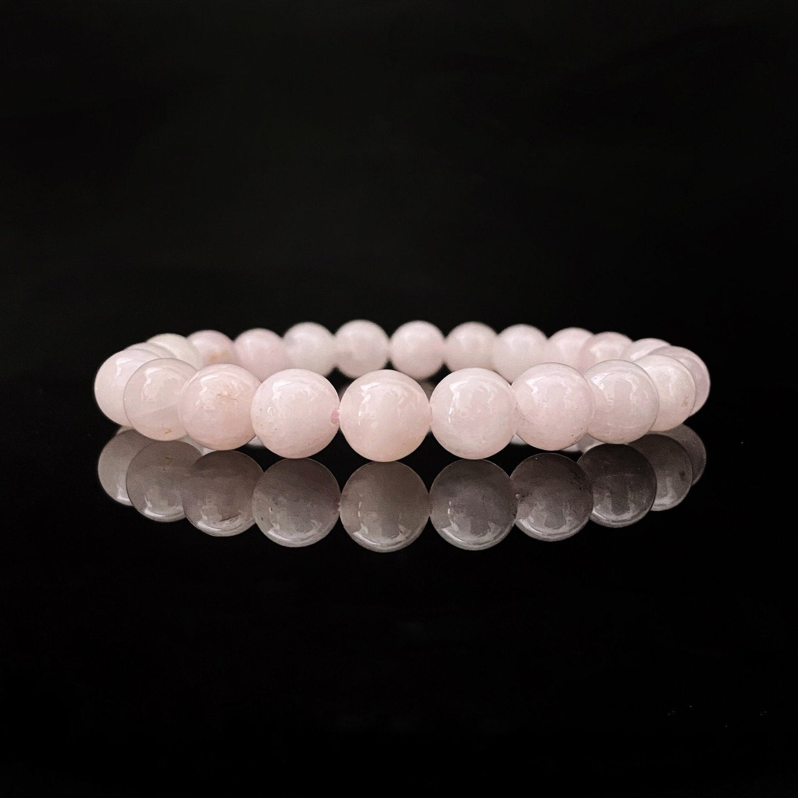 A delicate, light pink 8mm Rose Quartz Beaded Bracelet is showcased against a black background, with its reflection shimmering on the shiny surface below. The bracelet consists of smooth, round beads strung together in a continuous loop, perfect for balancing the Heart Chakra.
