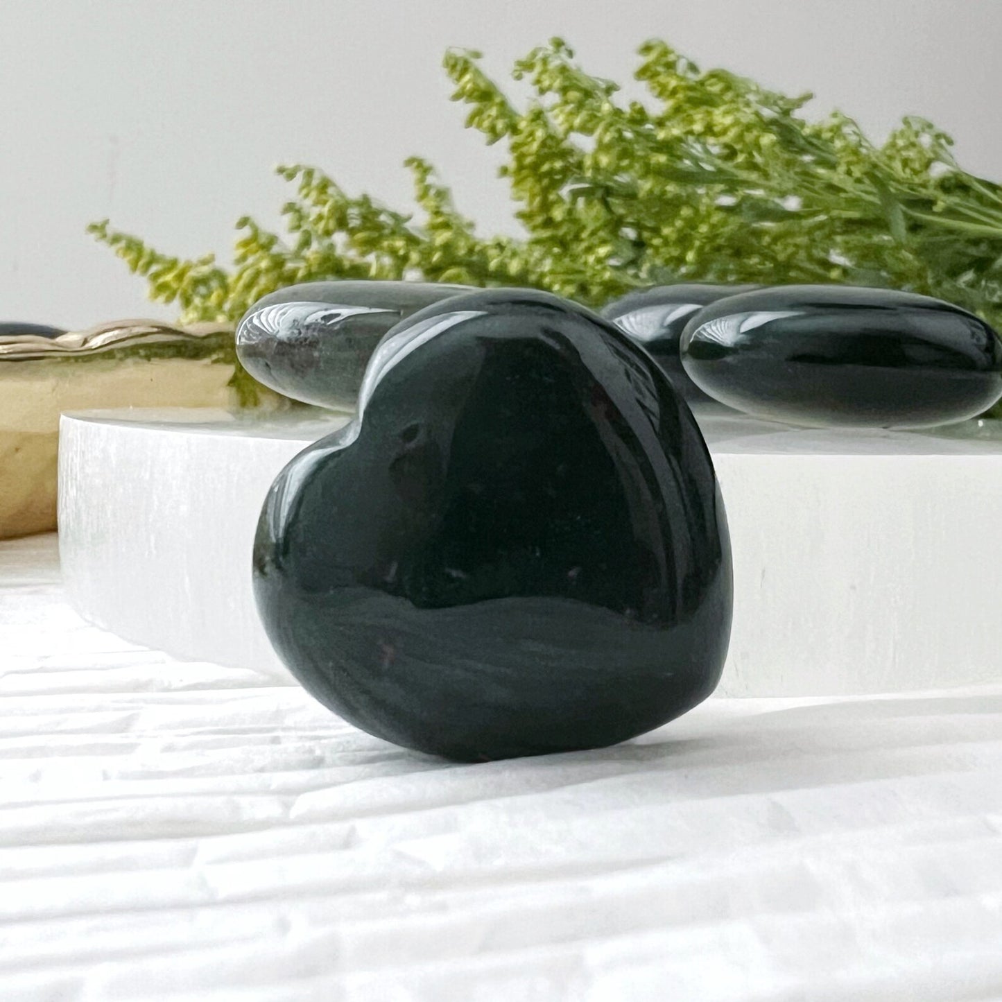 A set of small, polished 1" Mini Bloodstone Hearts displayed on a golden dish and a white circular platform. The background includes green foliage and a piece of paper with cursive text. These stones offer grounding and emotional support, with one heart-shaped stone placed beside the platform.