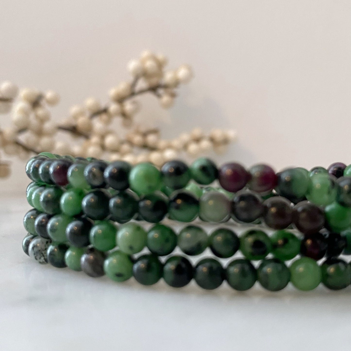 A close-up photo of a person wearing a 4mm Ruby Zoisite Beaded Bracelet on their wrist. The bracelet features polished round beads in varying shades of green, black, and a hint of purple. The background is slightly blurred with some out-of-focus decoration visible.