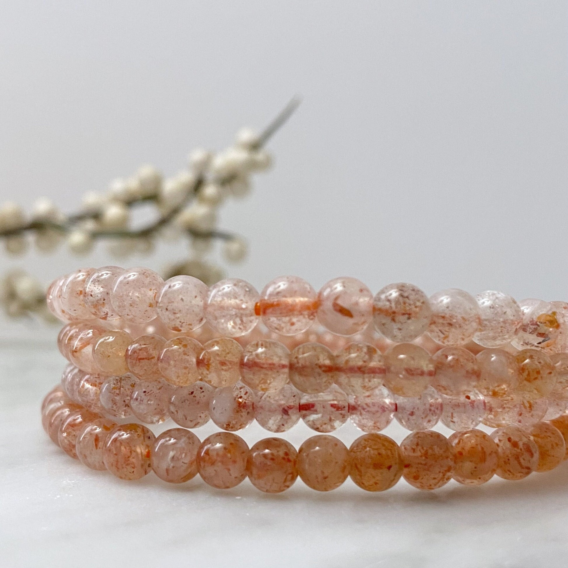 A round bracelet made of small, pale pink beads on a black background. The beads appear to be semi-translucent, with some subtle variations in color and texture. This 4mm Sunstone Beaded Bracelet is uniformly strung and neatly arranged in a circle, with a clear reflection below it.