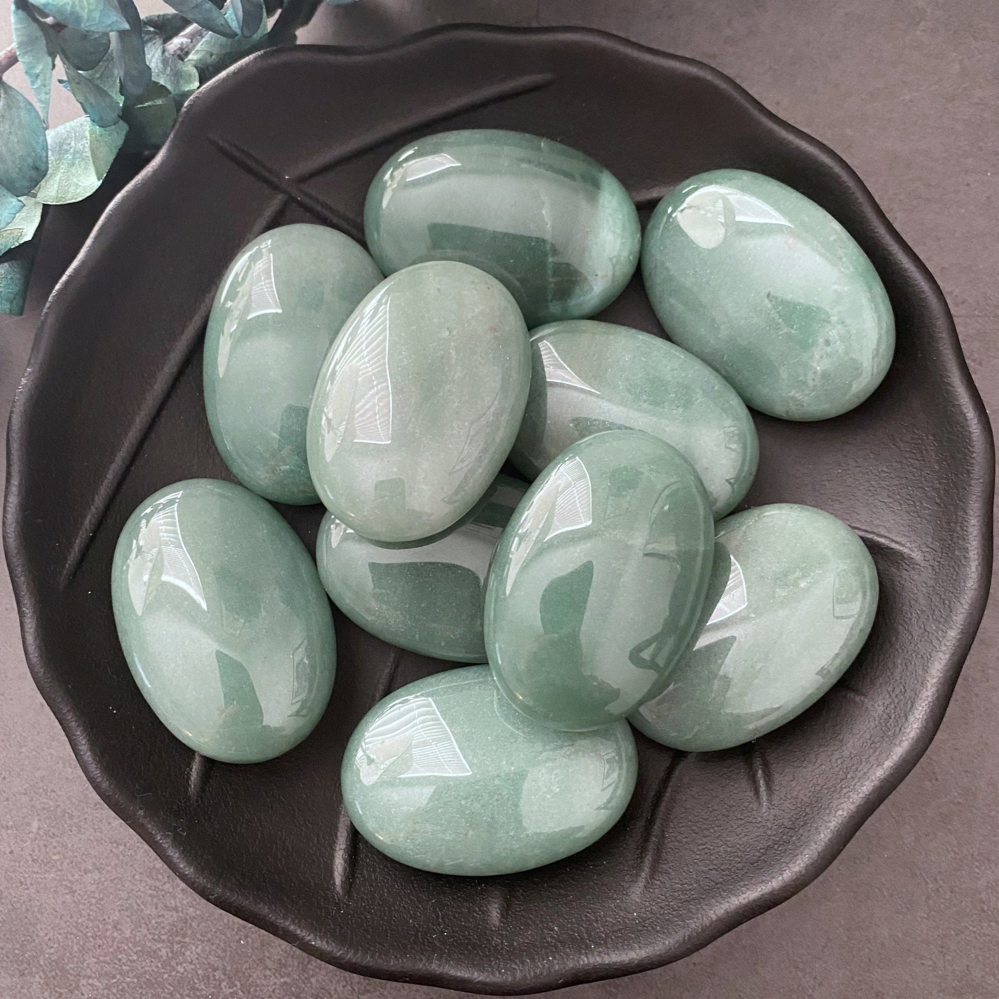 A black ceramic bowl filled with smooth, polished Green Aventurine Palm Stones is placed on a gray surface. Some green foliage decorates the top left corner of the image. The oval-shaped stones have a glossy finish and are perfect for meditation or as palm stones.