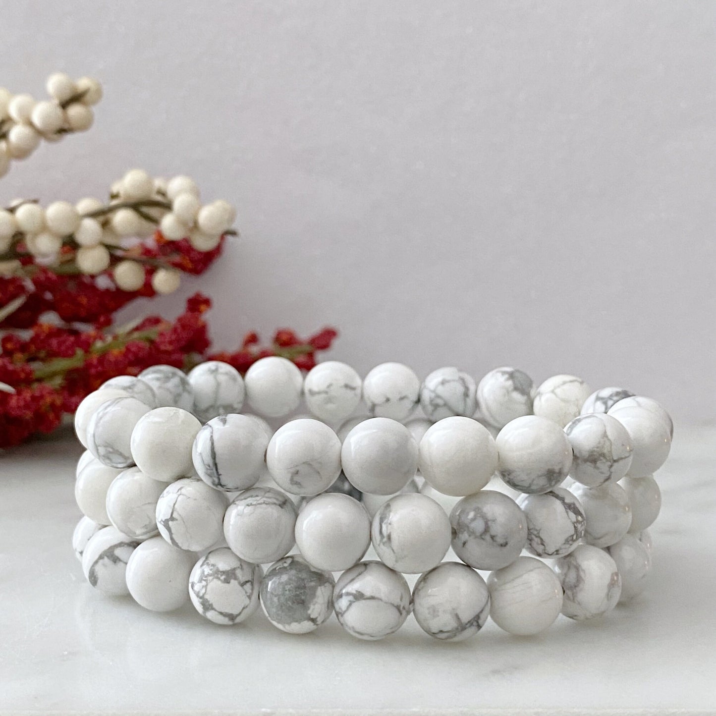 A stretchable 8mm Howlite Beaded Bracelet made of smooth, round white stones with light gray marbling, arranged in a circular pattern against a black background. The bracelet and its reflections create a symmetrical, elegant appearance that embodies the properties of an emotional healing stone.