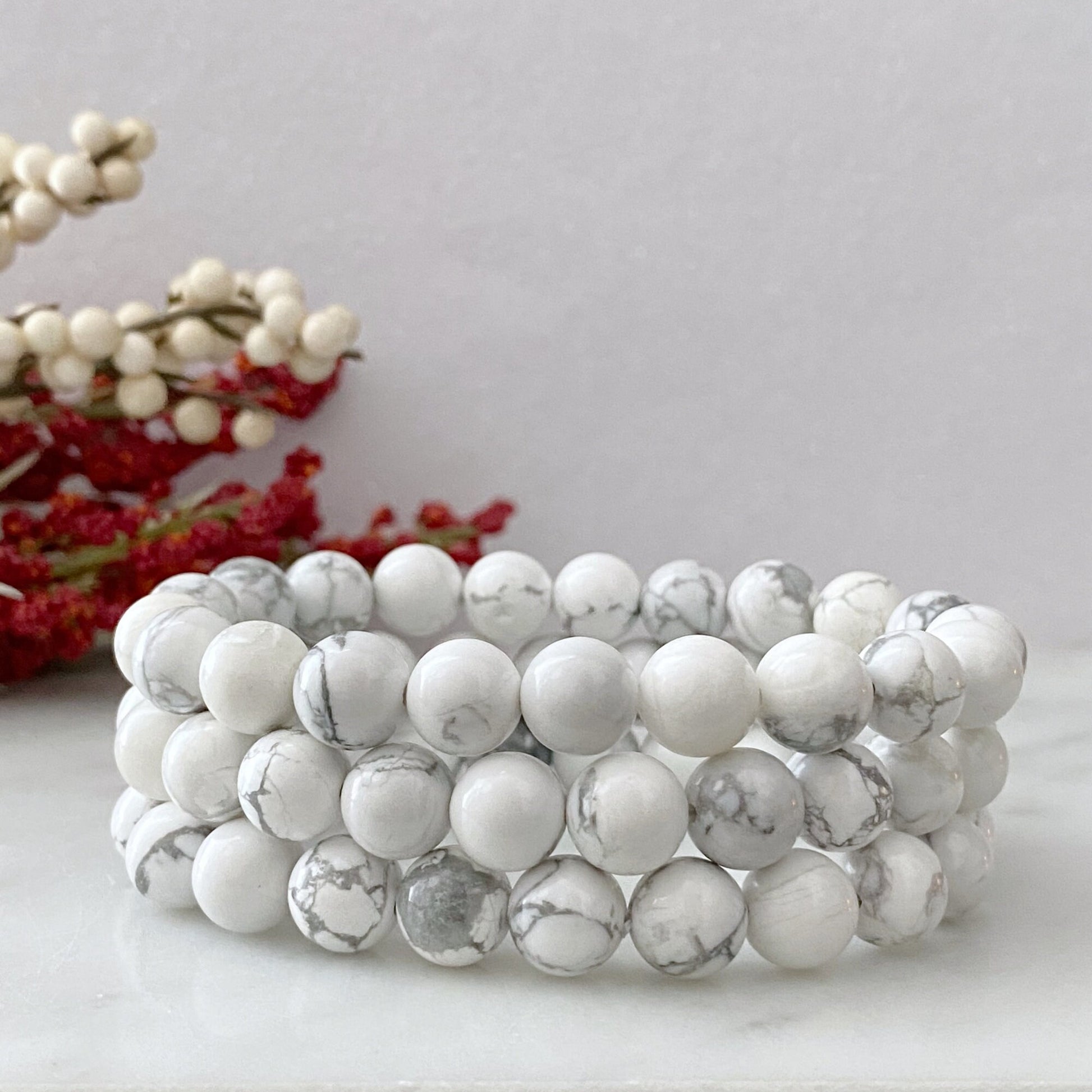 A stretchable 8mm Howlite Beaded Bracelet made of smooth, round white stones with light gray marbling, arranged in a circular pattern against a black background. The bracelet and its reflections create a symmetrical, elegant appearance that embodies the properties of an emotional healing stone.