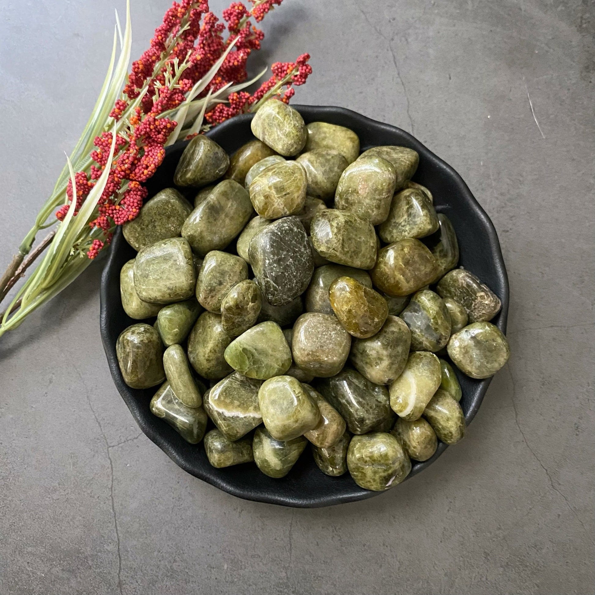 A black bowl filled with smooth Vesuvianite Tumbled Stones sits on a gray surface. Next to the bowl, there is a sprig of red berries with green leaves, adding a touch of color to the scene. These stones are known for promoting Heart Chakra balance and emotional healing.