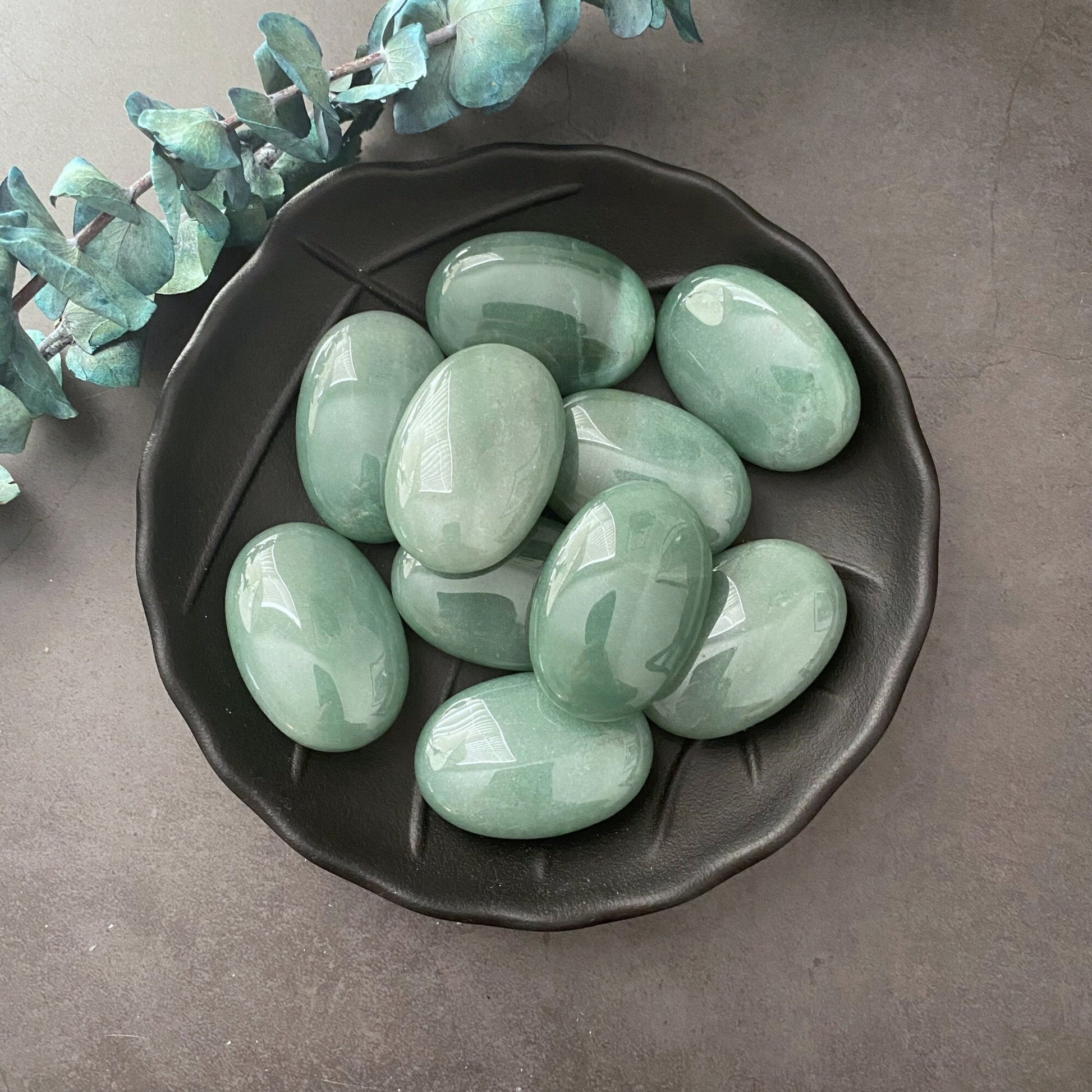 A black ceramic bowl filled with smooth, polished Green Aventurine Palm Stones is placed on a gray surface. Some green foliage decorates the top left corner of the image. The oval-shaped stones have a glossy finish and are perfect for meditation or as palm stones.