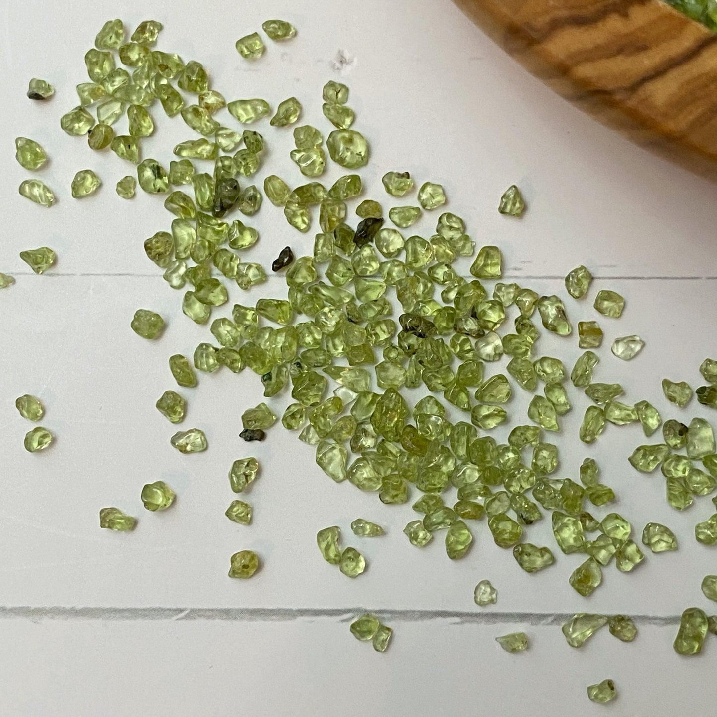 A wooden bowl filled with small green Peridot Crystal Chips, with some spilled on the white surface. Beside the bowl are two small vials containing similar crystals, a smudge stick for metaphysical uses, and a small plastic bag also containing the green crystals.