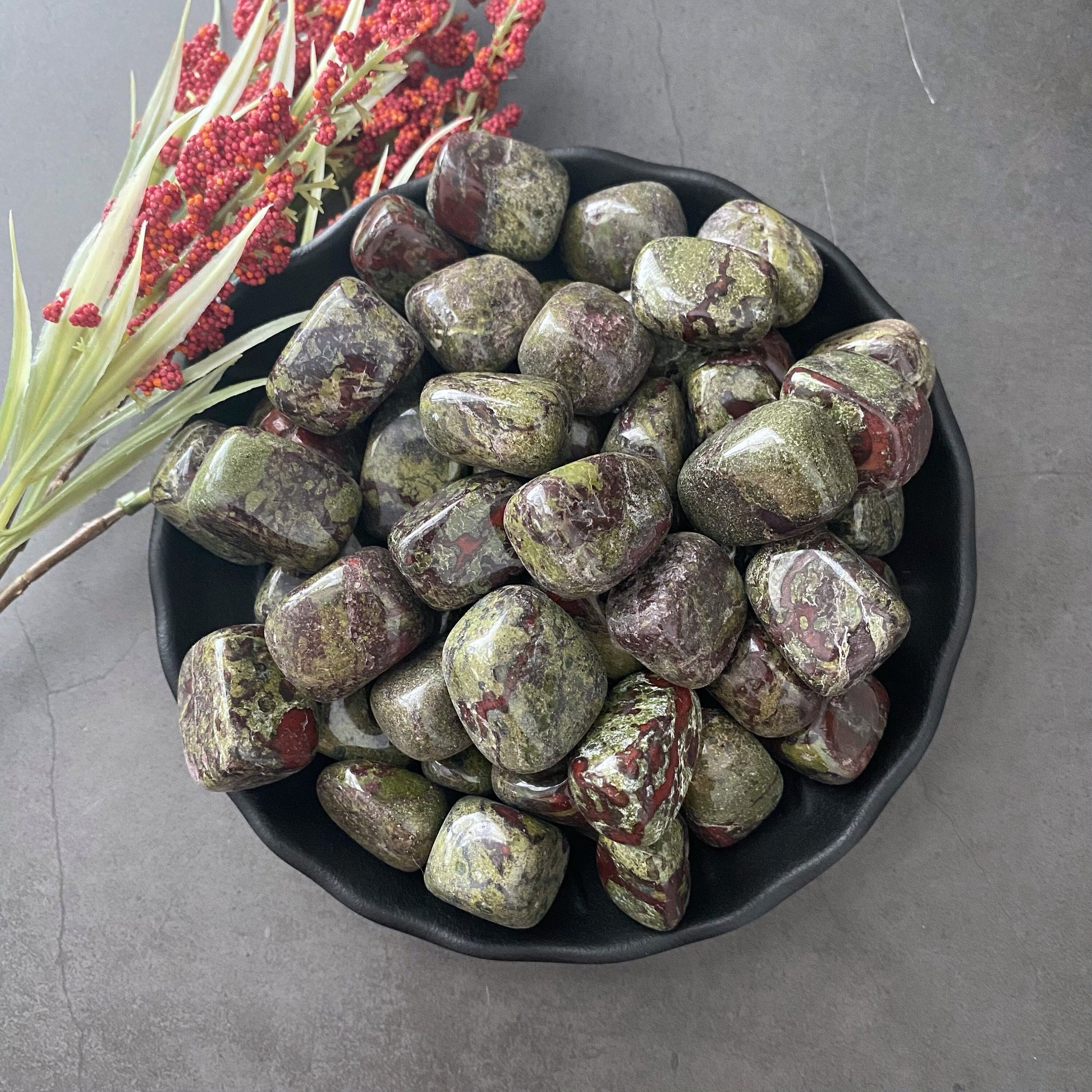 A black bowl filled with polished tumbled stones, including Heart Chakra Stones and Dragon Blood Jasper Tumbled Stones, varying in shades of green and purple, displayed on a gray surface. To the left of the bowl, a sprig of red berries and green foliage is visible.