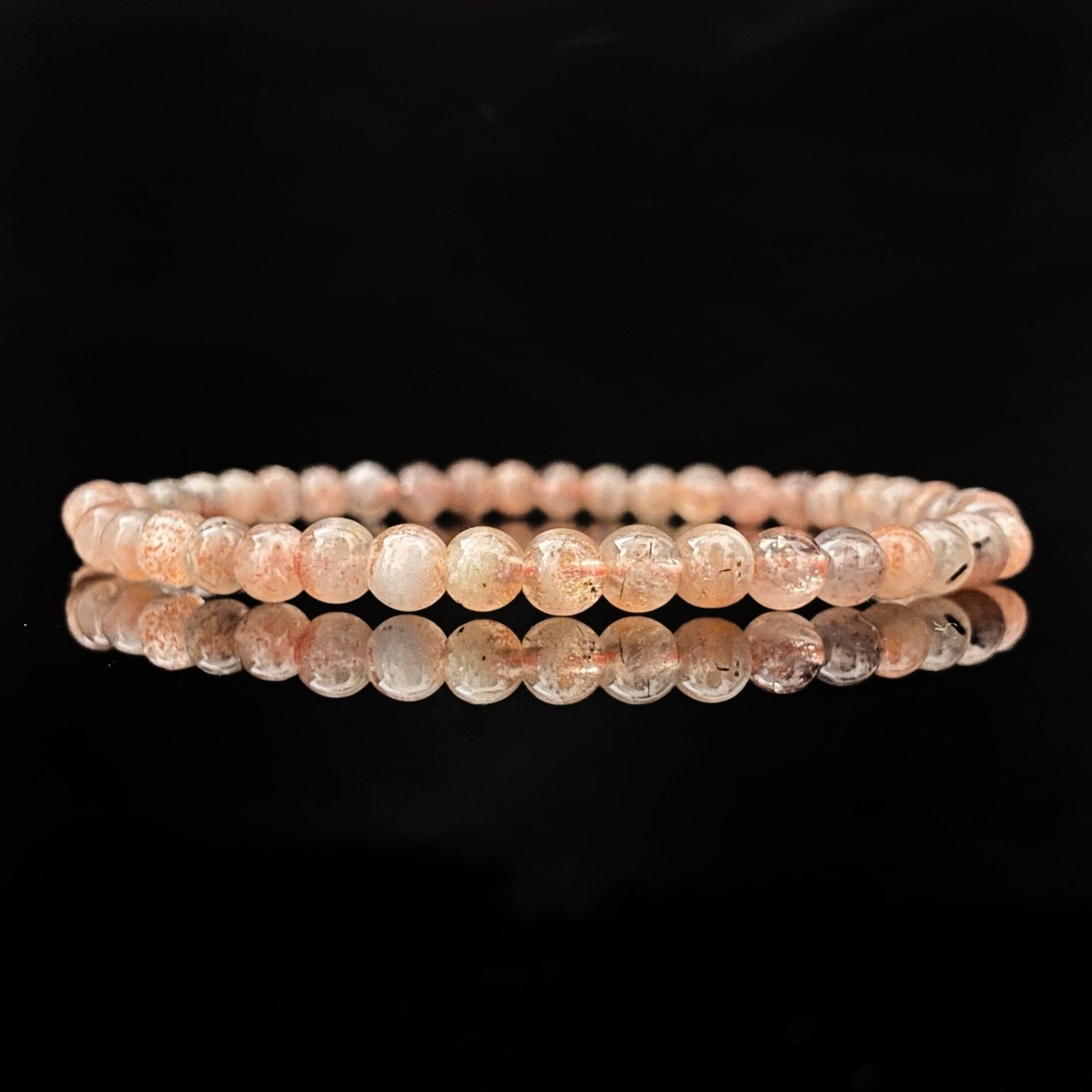 A round bracelet made of small, pale pink beads on a black background. The beads appear to be semi-translucent, with some subtle variations in color and texture. This 4mm Sunstone Beaded Bracelet is uniformly strung and neatly arranged in a circle, with a clear reflection below it.