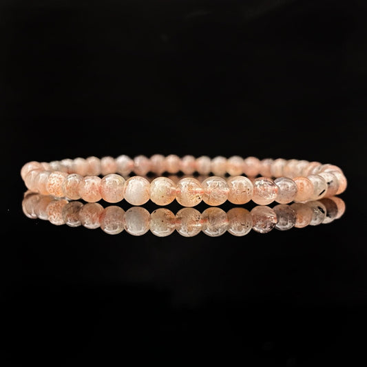 A round bracelet made of small, pale pink beads on a black background. The beads appear to be semi-translucent, with some subtle variations in color and texture. This 4mm Sunstone Beaded Bracelet is uniformly strung and neatly arranged in a circle, with a clear reflection below it.
