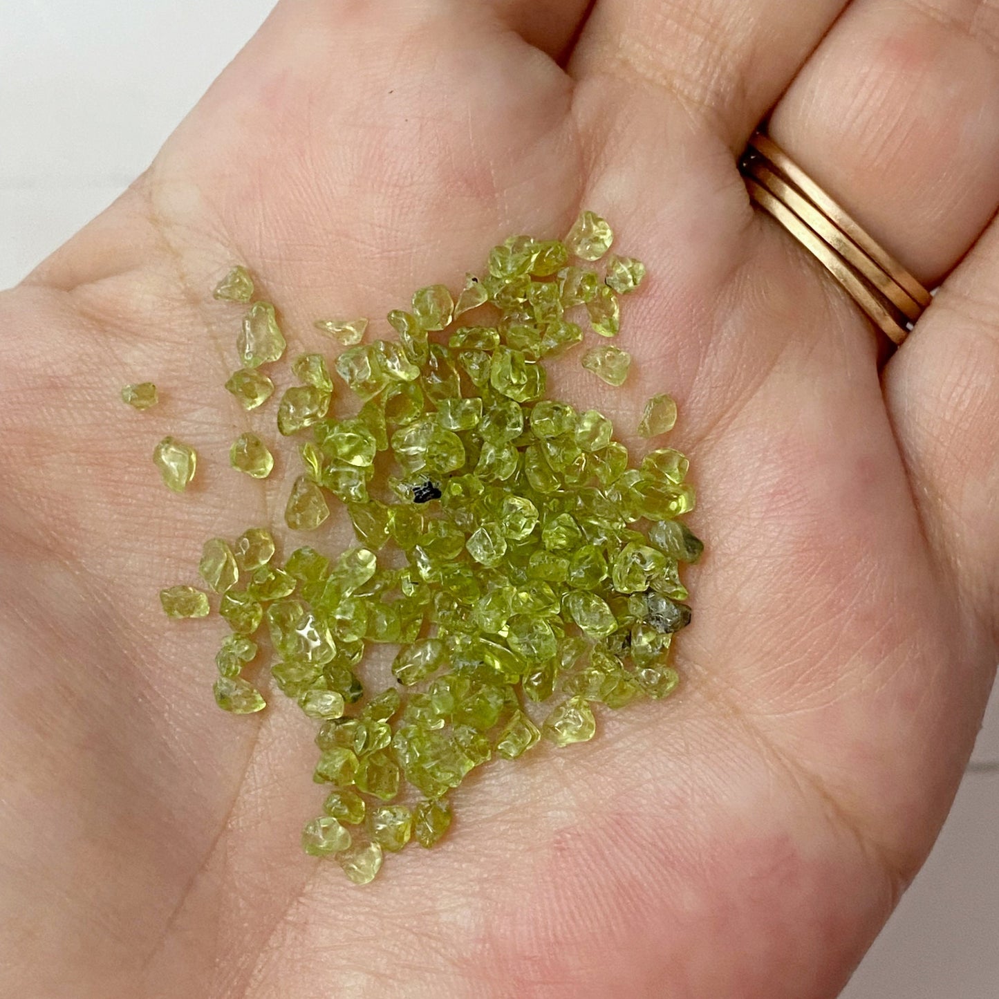 A wooden bowl filled with small green Peridot Crystal Chips, with some spilled on the white surface. Beside the bowl are two small vials containing similar crystals, a smudge stick for metaphysical uses, and a small plastic bag also containing the green crystals.