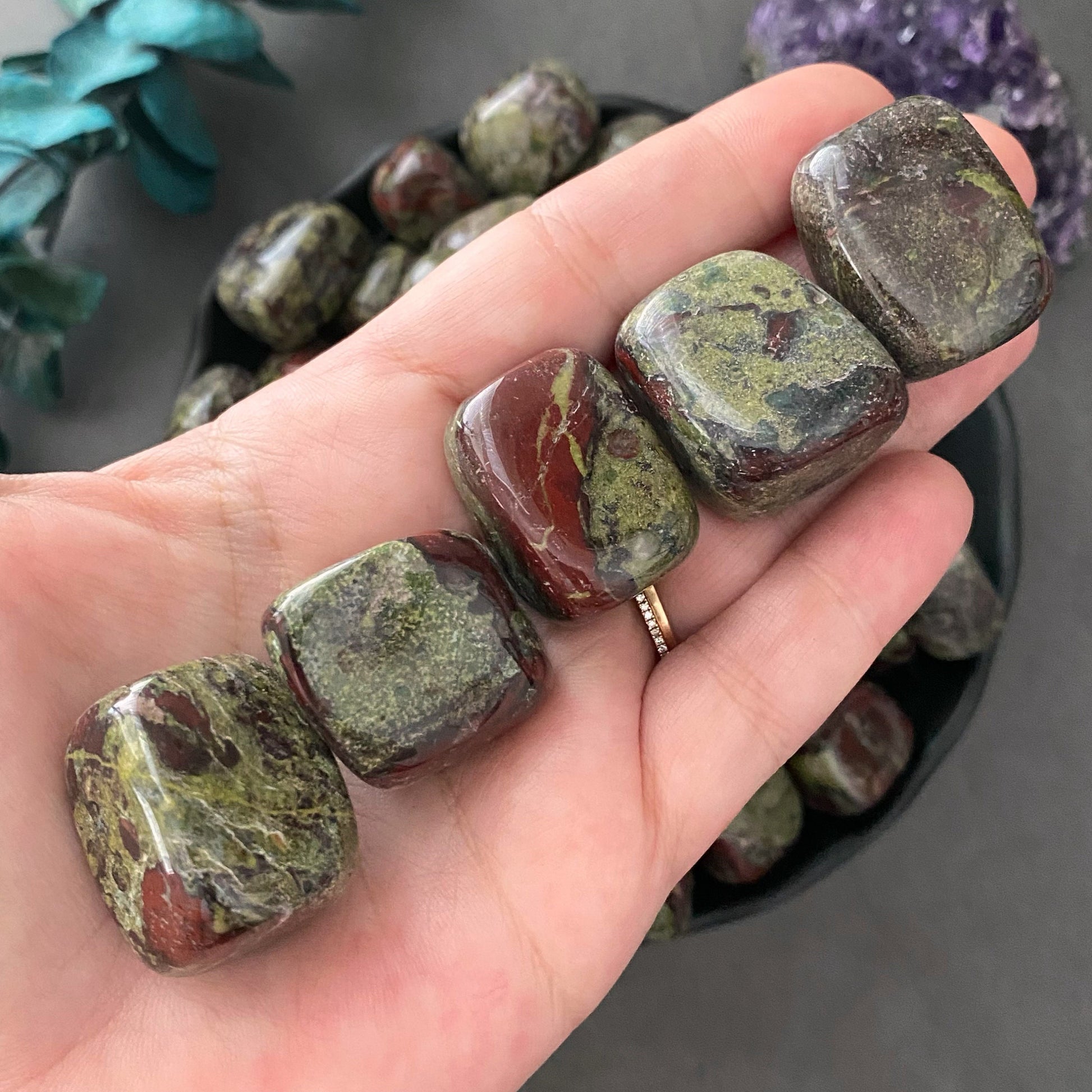 A black bowl filled with polished tumbled stones, including Heart Chakra Stones and Dragon Blood Jasper Tumbled Stones, varying in shades of green and purple, displayed on a gray surface. To the left of the bowl, a sprig of red berries and green foliage is visible.