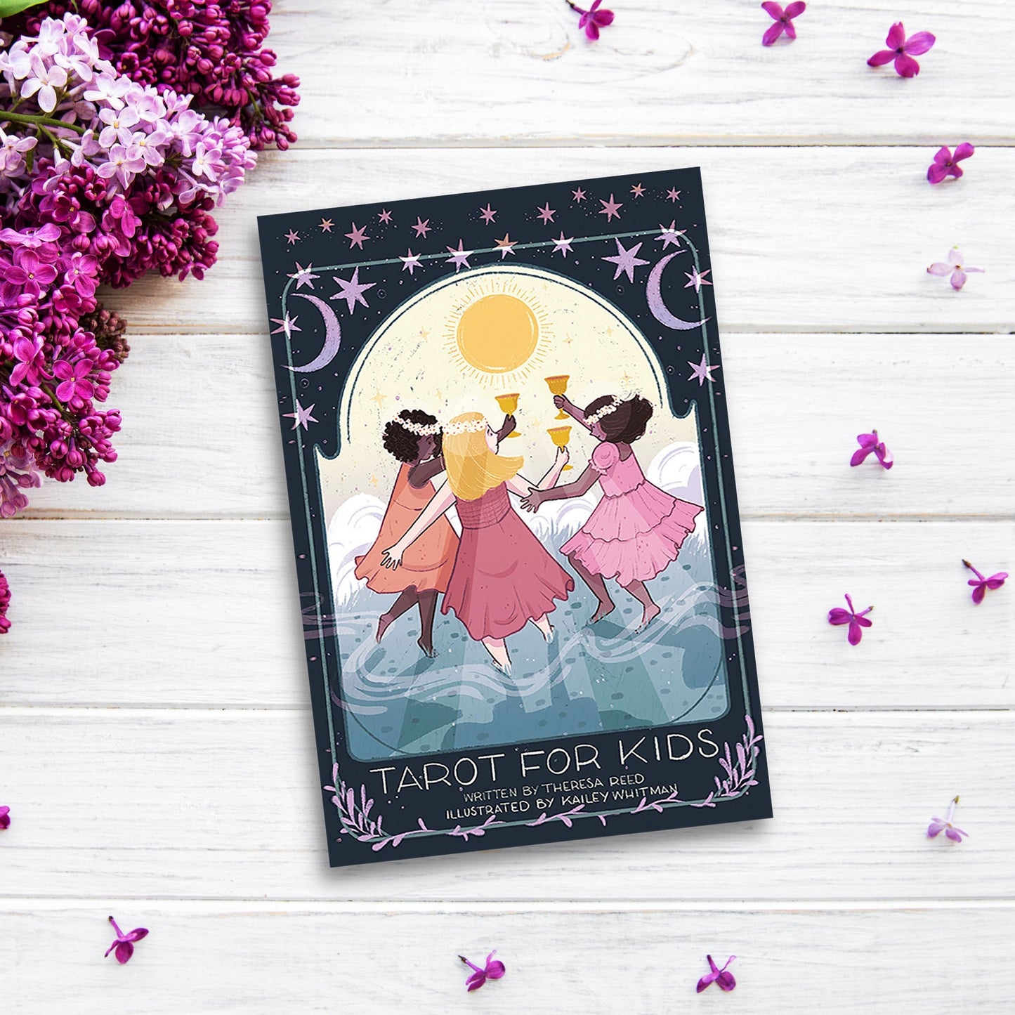 A children card deck titled "Tarot for Kids" is placed on a white wooden surface with scattered purple flowers and lilac blossoms around. The cover features three children in pink dresses dancing under a yellow sun, with a dark blue sky and celestial designs, perfect for enhancing decision-making skills.