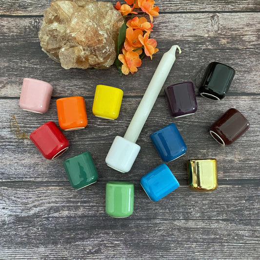 A white candle stands upright, surrounded by Ceramic Candle Holders for Chime Ritual Candles (14 Colors), including shades of pink, orange, yellow, green, blue, black, red, and a metallic gold. An orange flower and a large rock add natural elements on a wooden surface.