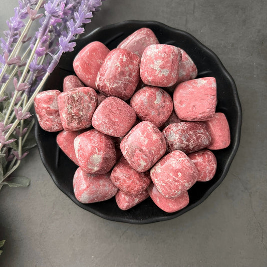 A black bowl filled with polished, pinkish-red Thulite Tumbled Stones, a renowned Heart Chakra stone, sits on a dark surface. Sprigs of light purple lavender are placed to the left of the bowl, adding a touch of natural beauty and emotional healing to the scene.