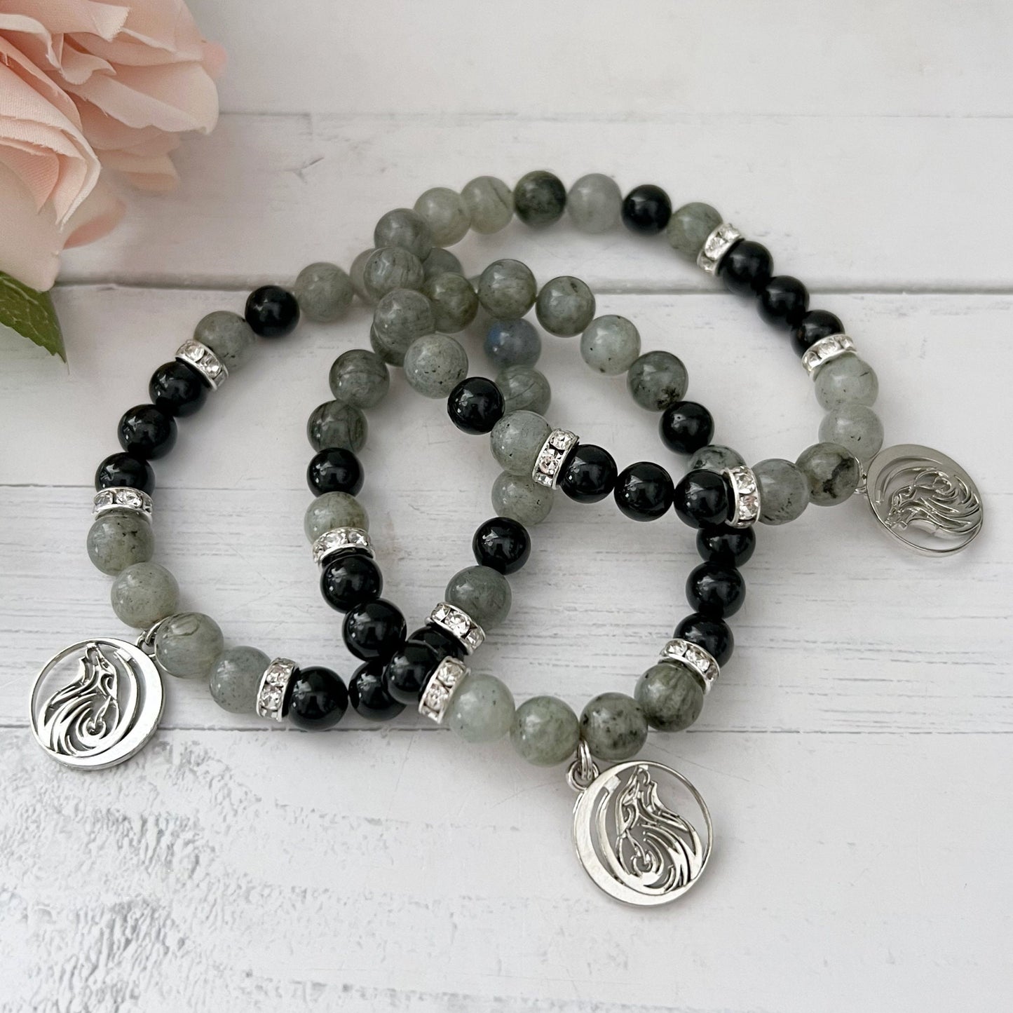 An 8mm Labradorite & Black Tourmaline Beaded Bracelet w/ Wolf Charm is displayed on a wooden surface. The bracelet features silver spacers and a silver pendant with an intricate design. Pink rose petals are partially visible in the background.