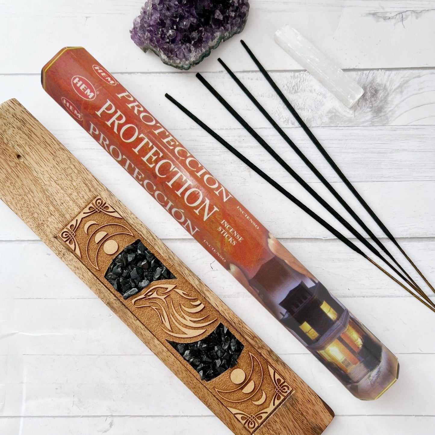 A rectangular box and a cylindrical tube of Protection Incense Sticks. The packaging features an image of a house being shielded by two hands. The text on the box reads "Protection Incense Sticks." Perfect for enhancing your meditation or yoga sessions, the product is handcrafted in India.