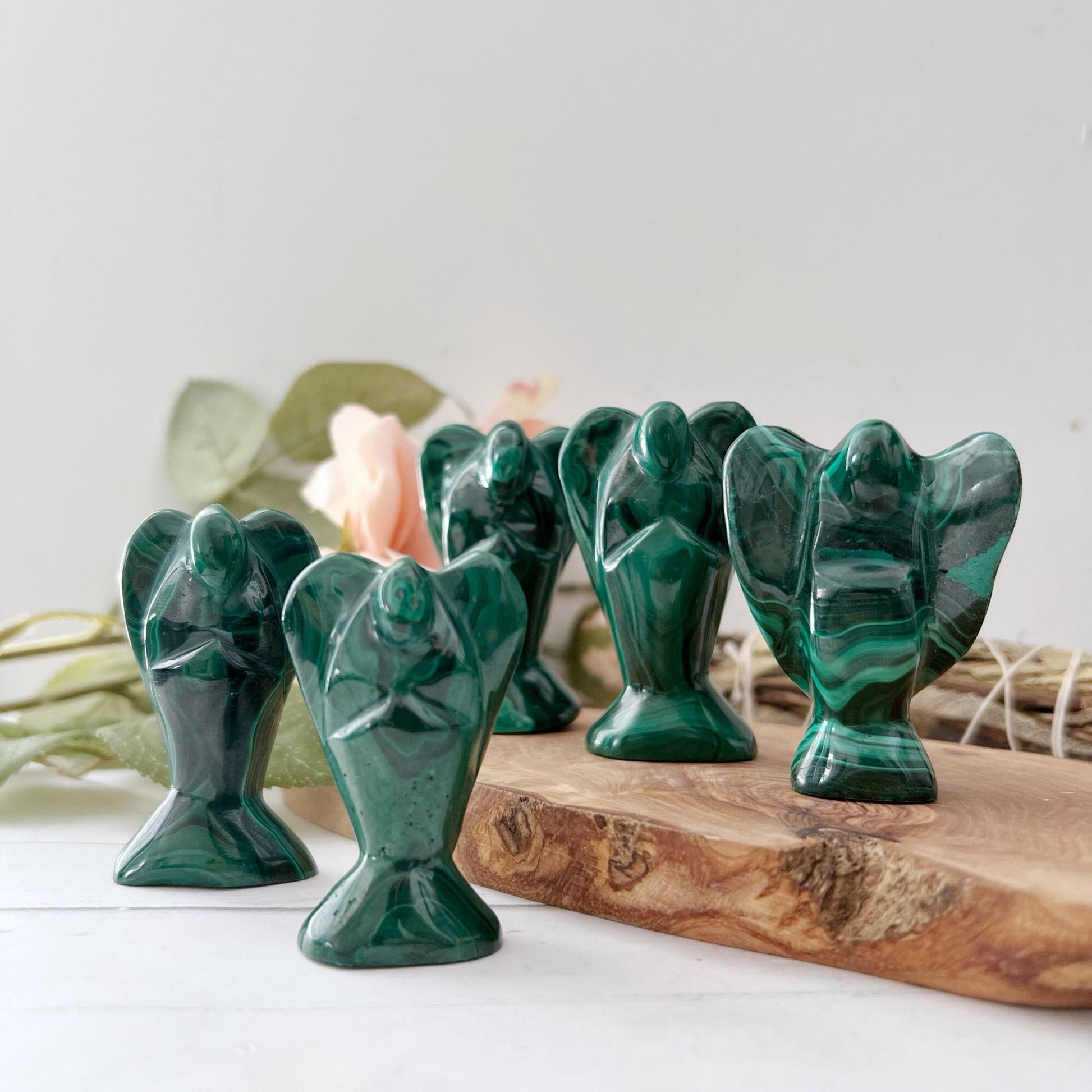 2" Genuine Malachite Angels are arranged on a wooden slab. Each hand-carved figurine is distinct, showcasing the stone’s unique patterns and deep green hues. The background is softly lit with out-of-focus leaves and a pale flower, adding a serene and natural touch.