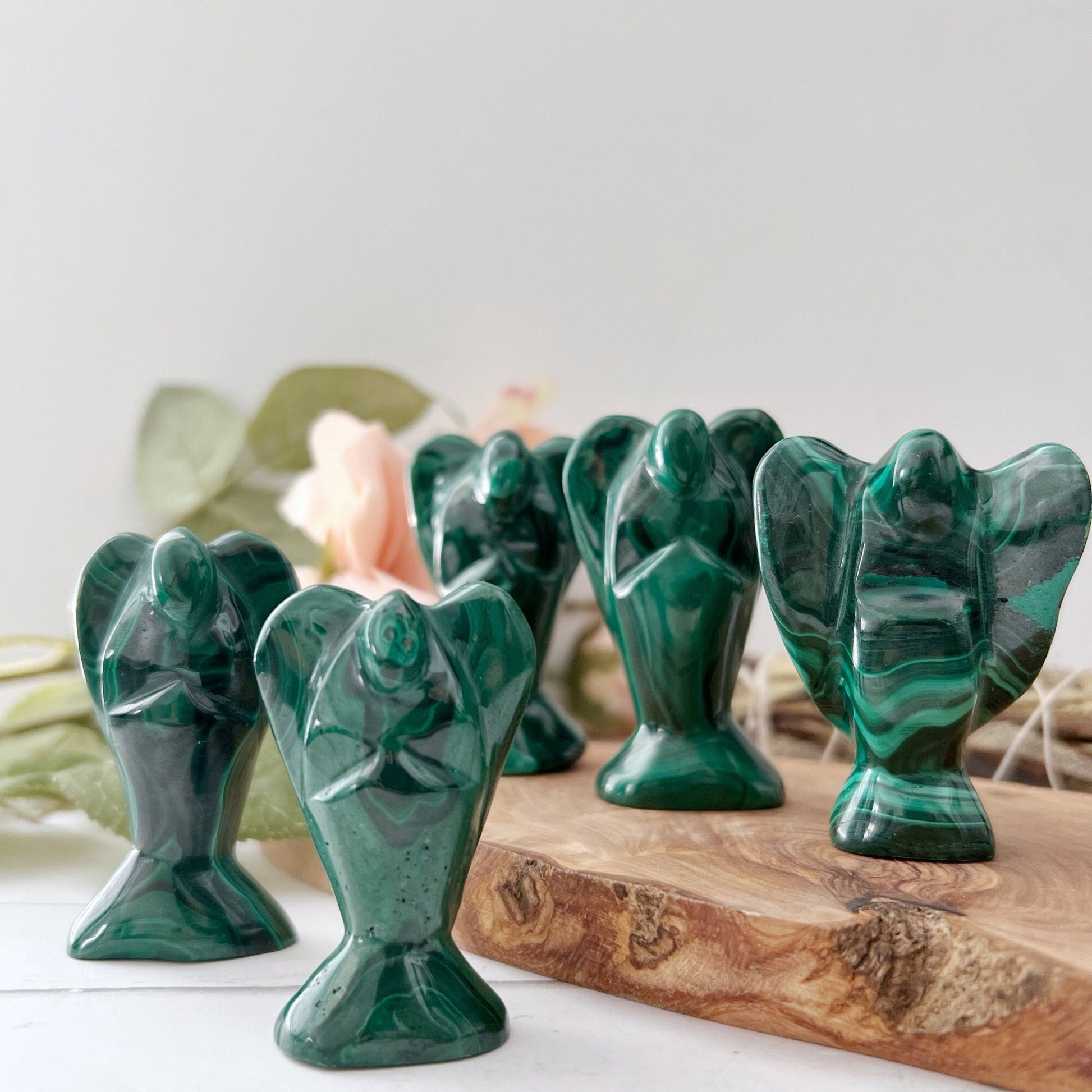 2" Genuine Malachite Angels are arranged on a wooden slab. Each hand-carved figurine is distinct, showcasing the stone’s unique patterns and deep green hues. The background is softly lit with out-of-focus leaves and a pale flower, adding a serene and natural touch.