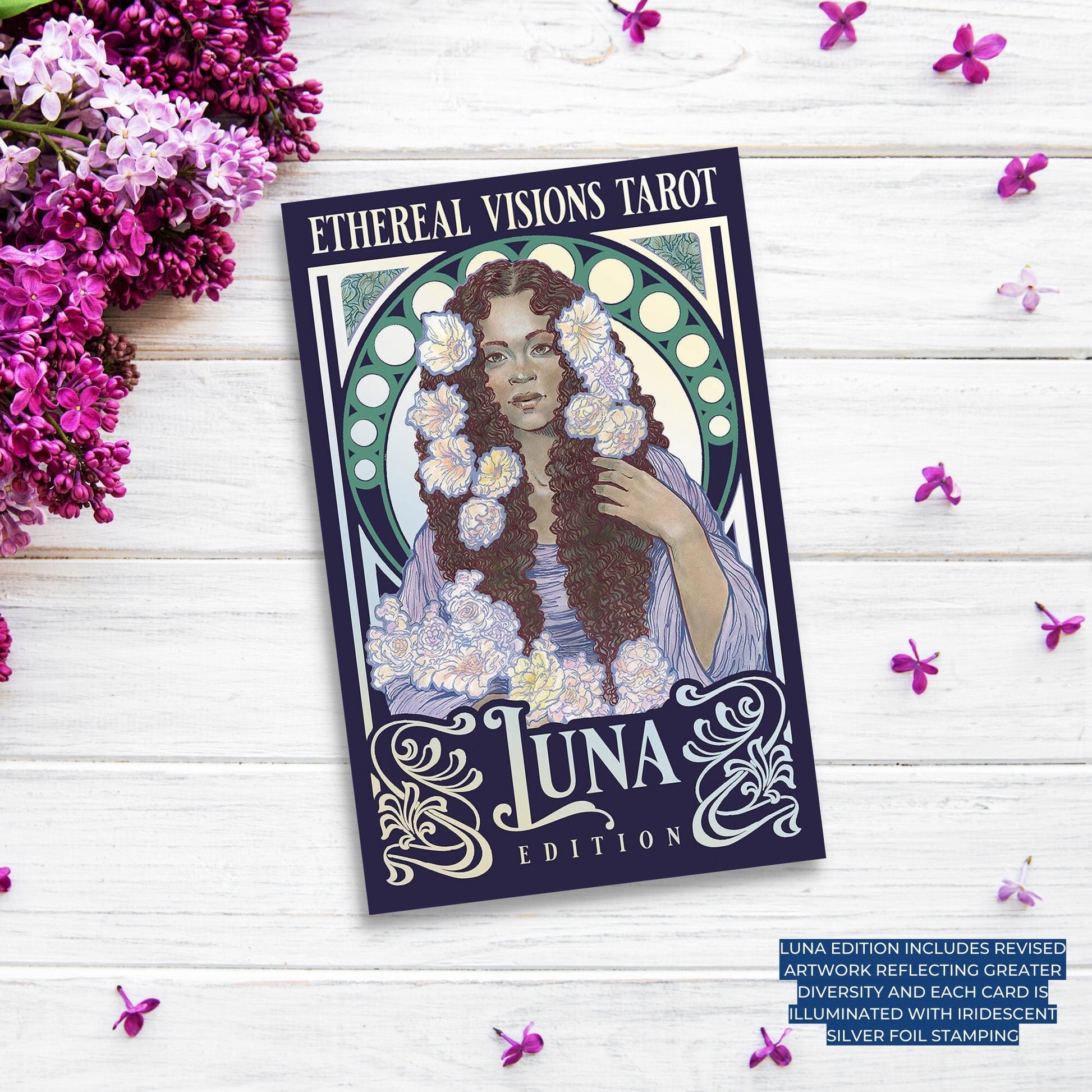 A box of the "Ethereal Visions Luna Edition Tarot" is placed on a light wooden surface surrounded by purple and white flowers. The Art Nouveau-style box features an illustration of a woman with flowers in her hair and around her neck, accented by a decorative border and circular design.