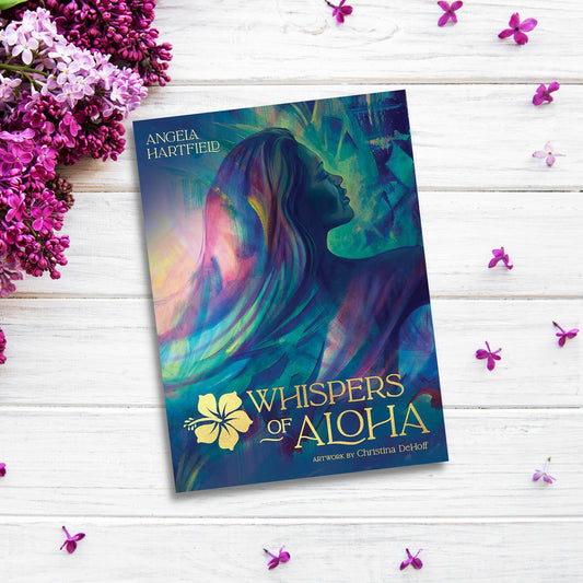 A book titled "Whispers of Aloha Oracle Deck" by Angela Hartfield, featuring artwork by Christina DeHoff, is placed on a white wooden surface adorned with purple flowers and petals. Reminiscent of Hawaii, the cover art shows a colorful silhouette of a woman with flowing hair.