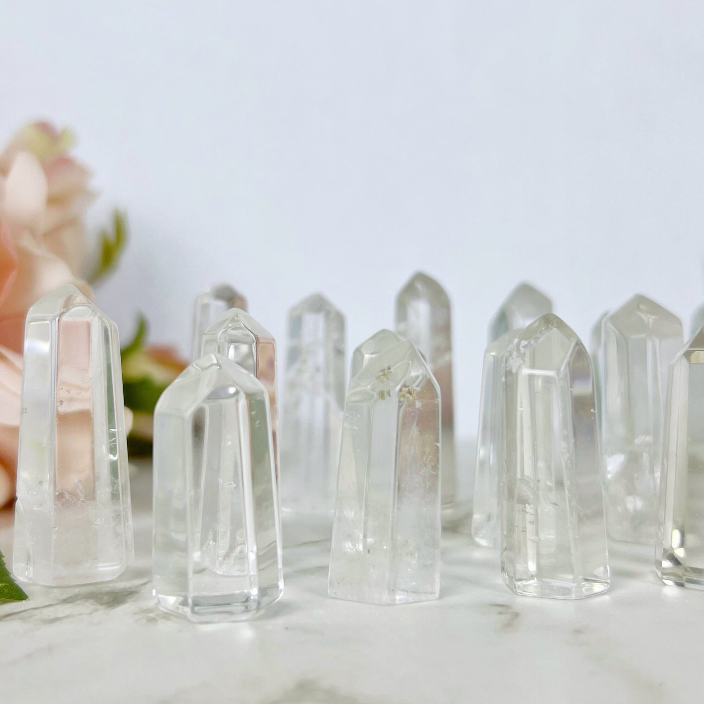 A wooden bowl filled with multiple 1-2" Mini Clear Quartz Points, known as the master healer, sits on a marble surface. A pale pink rose with green leaves lies next to the bowl, adding a touch of softness to the natural and serene composition.