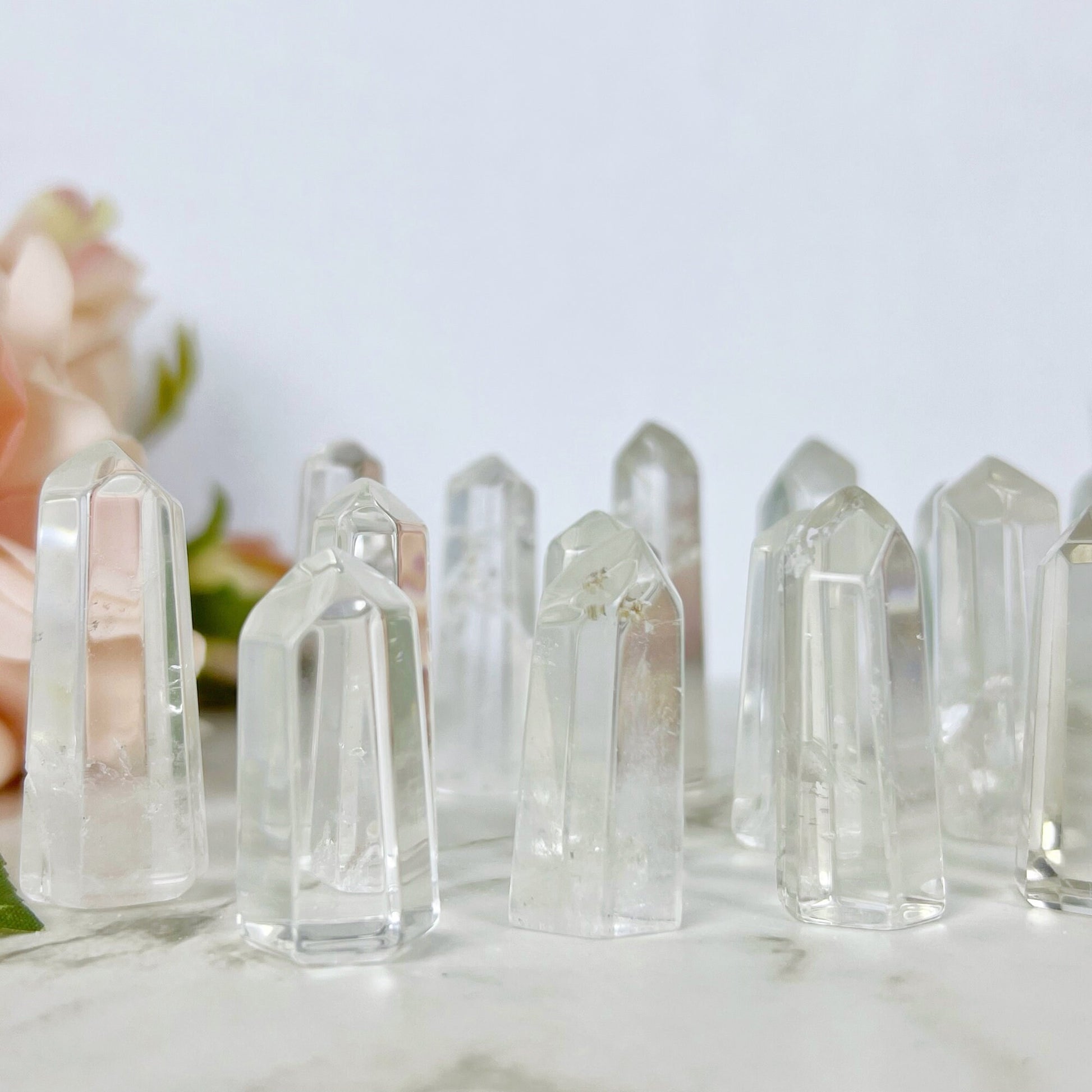 A wooden bowl filled with multiple 1-2" Mini Clear Quartz Points, known as the master healer, sits on a marble surface. A pale pink rose with green leaves lies next to the bowl, adding a touch of softness to the natural and serene composition.