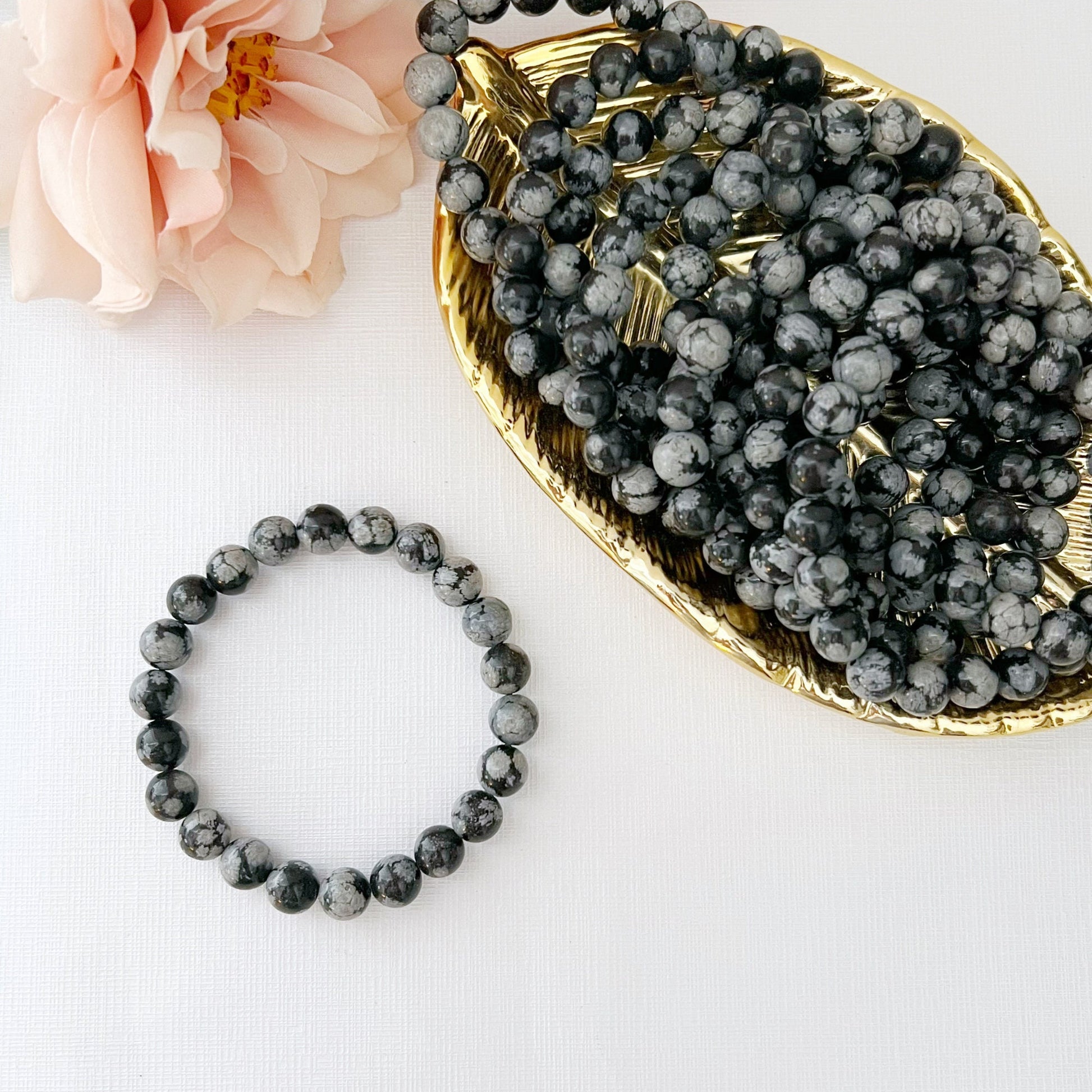 A delicate 8mm Snowflake Obsidian Beaded Bracelet made of round black and gray speckled beads is displayed against a white background. Nearby, there is a soft-focus flower and a golden dish filled with similar beads, radiating healing energy.