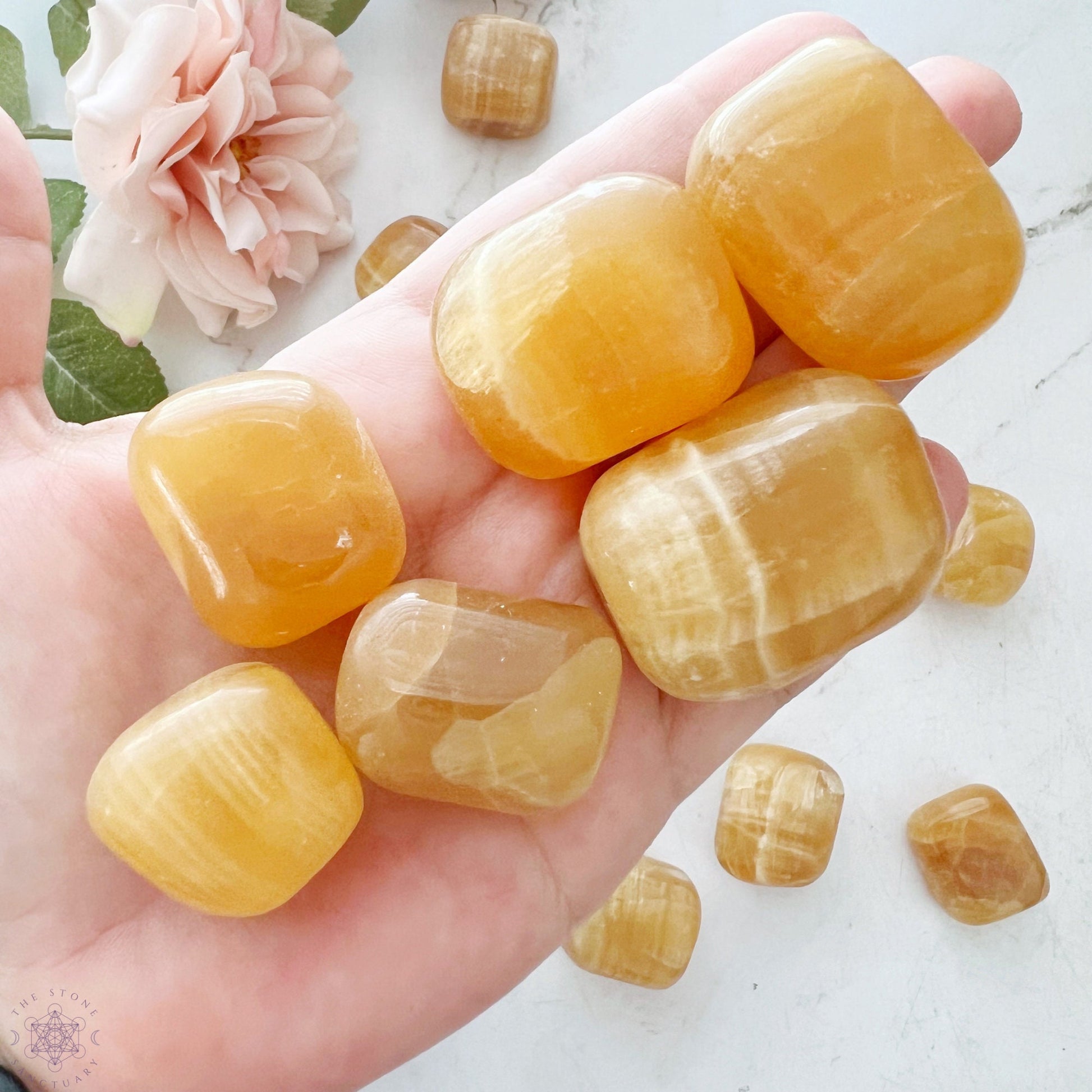 A round wooden bowl filled with polished, yellow-orange Honey Calcite Tumbled Stones. The tumbled stones have a smooth, glossy surface and vary in size and shape, giving the bowl a vibrant and reflective appearance. The white background contrasts with the bowl’s natural tones.