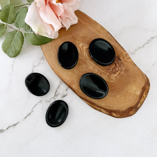 A wooden tray on a marble surface holds five polished black stones of different sizes. Among them, an Obsidian Worry Stone stands out as a protective gemstone believed to aid in emotional healing. A light pink rose with green leaves is placed next to the tray.
Notice that the product name "Obsidian Worry Stone" already appears correctly in the sentence.
