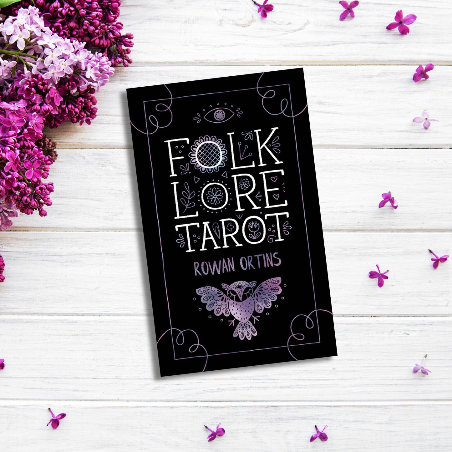 A black tarot card deck box titled "Folklore Tarot" by Rowan Ortins lies on a white wooden surface surrounded by purple and lilac flowers. The box features decorative white and purple designs, including an eye, floral patterns, and a bird. It comes with an illustrated guidebook exploring mythological traditions.