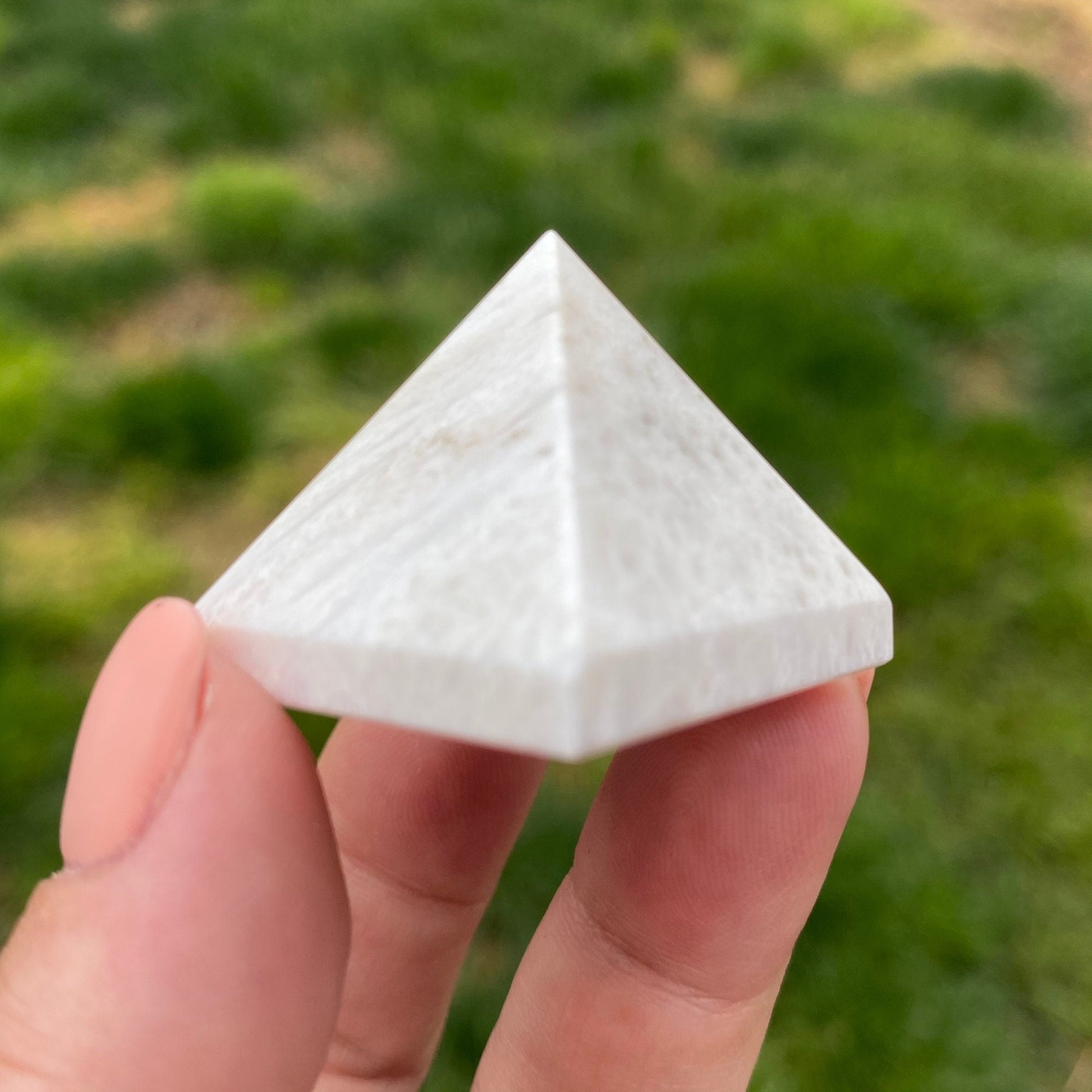 A hand holding a small, white 1" Mini Scolecite Pyramid outdoors with grass in the background, perfect as a meditation tool for achieving inner peace.