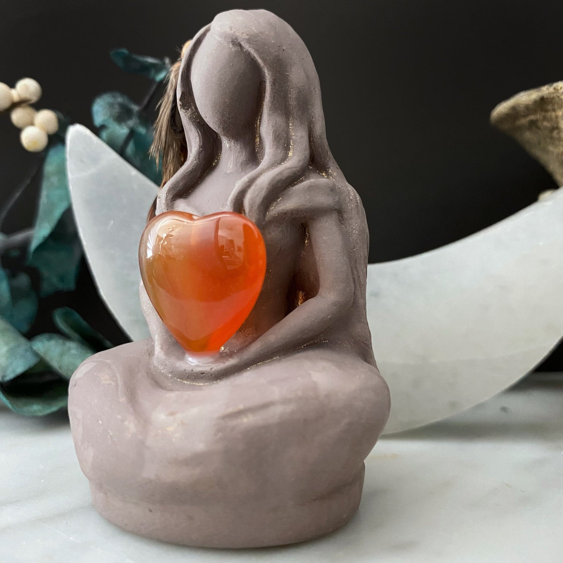 A serene, faceless Aine Goddess Statue sits cross-legged, holding an orange heart-shaped gem. The Celtic Goddess statue features feather details on its side and is surrounded by greenery and small decorative elements, including a large crescent moon shape in the background.