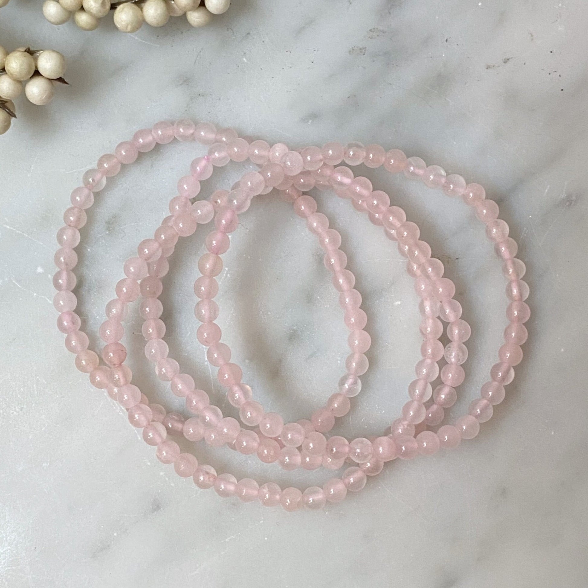 A close-up image shows a 4mm Rose Quartz Beaded Bracelet made of semi-precious stones, coiled in a circular pattern. The backdrop features an out-of-focus branch adorned with small white berries, set against a light, soft-focus background, enhancing the bracelet’s connection to Heart Chakra healing.