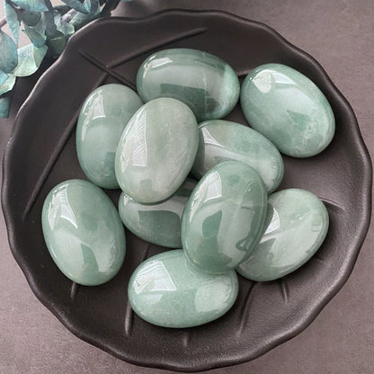 A black ceramic bowl filled with smooth, polished Green Aventurine Palm Stones is placed on a gray surface. Some green foliage decorates the top left corner of the image. The oval-shaped stones have a glossy finish and are perfect for meditation or as palm stones.

