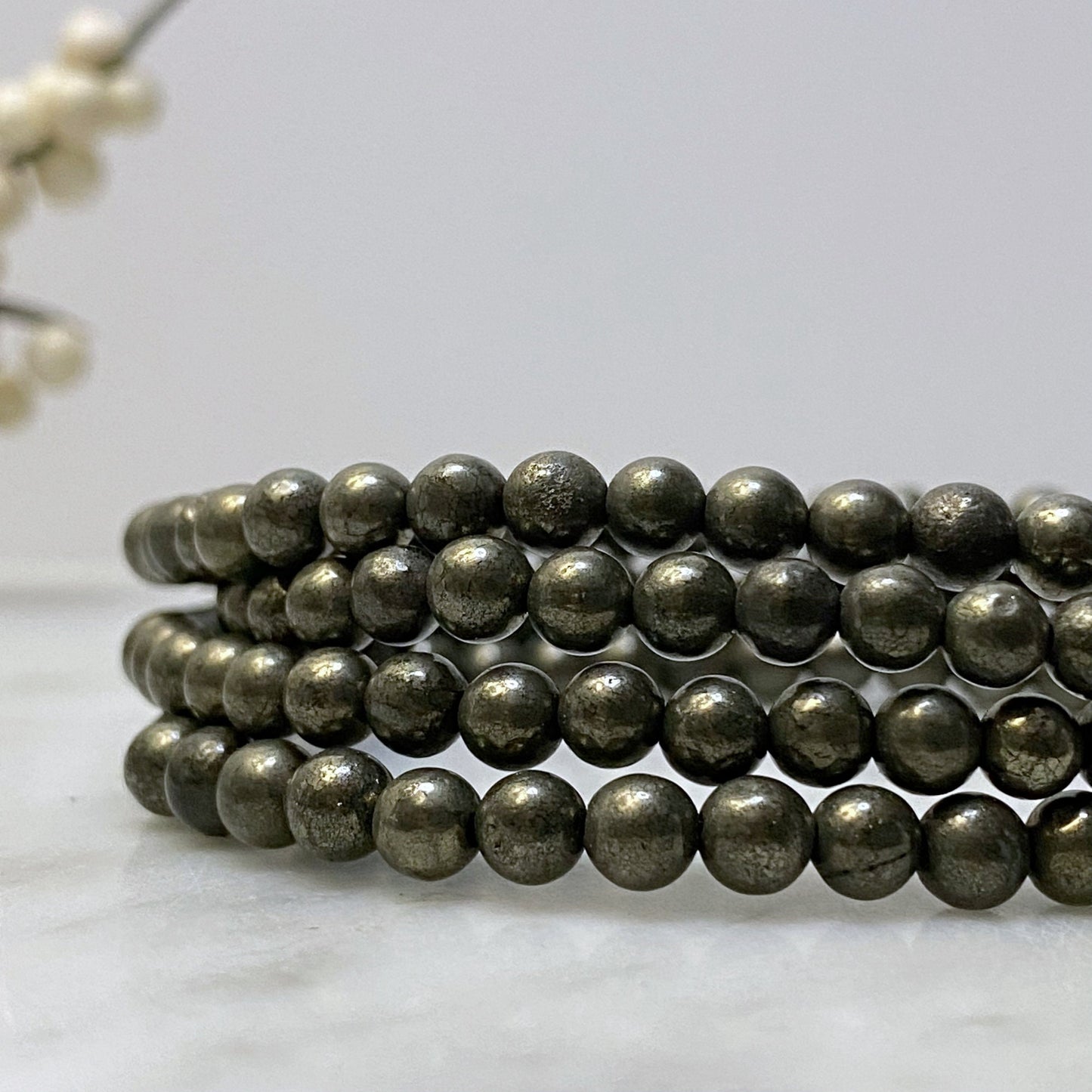 A close-up image of a 4mm Pyrite Beaded Bracelet made of metallic spherical beads, coiled neatly in layers. The shiny, reflective beads suggest it's a protection stone. The background is minimalist, with a blurred decorative element.