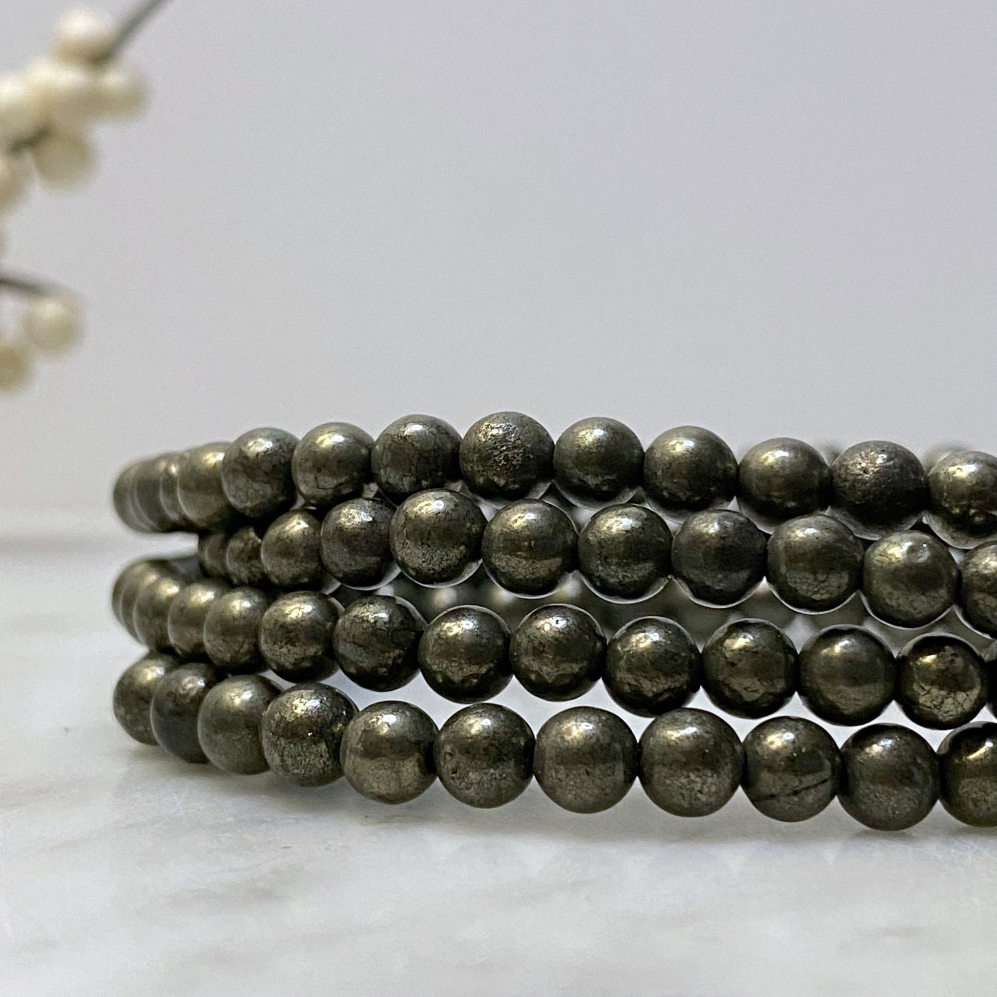 A close-up image of a 4mm Pyrite Beaded Bracelet made of metallic spherical beads, coiled neatly in layers. The shiny, reflective beads suggest it's a protection stone. The background is minimalist, with a blurred decorative element.
