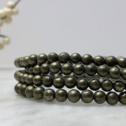 A close-up image of a 4mm Pyrite Beaded Bracelet made of metallic spherical beads, coiled neatly in layers. The shiny, reflective beads suggest it's a protection stone. The background is minimalist, with a blurred decorative element.