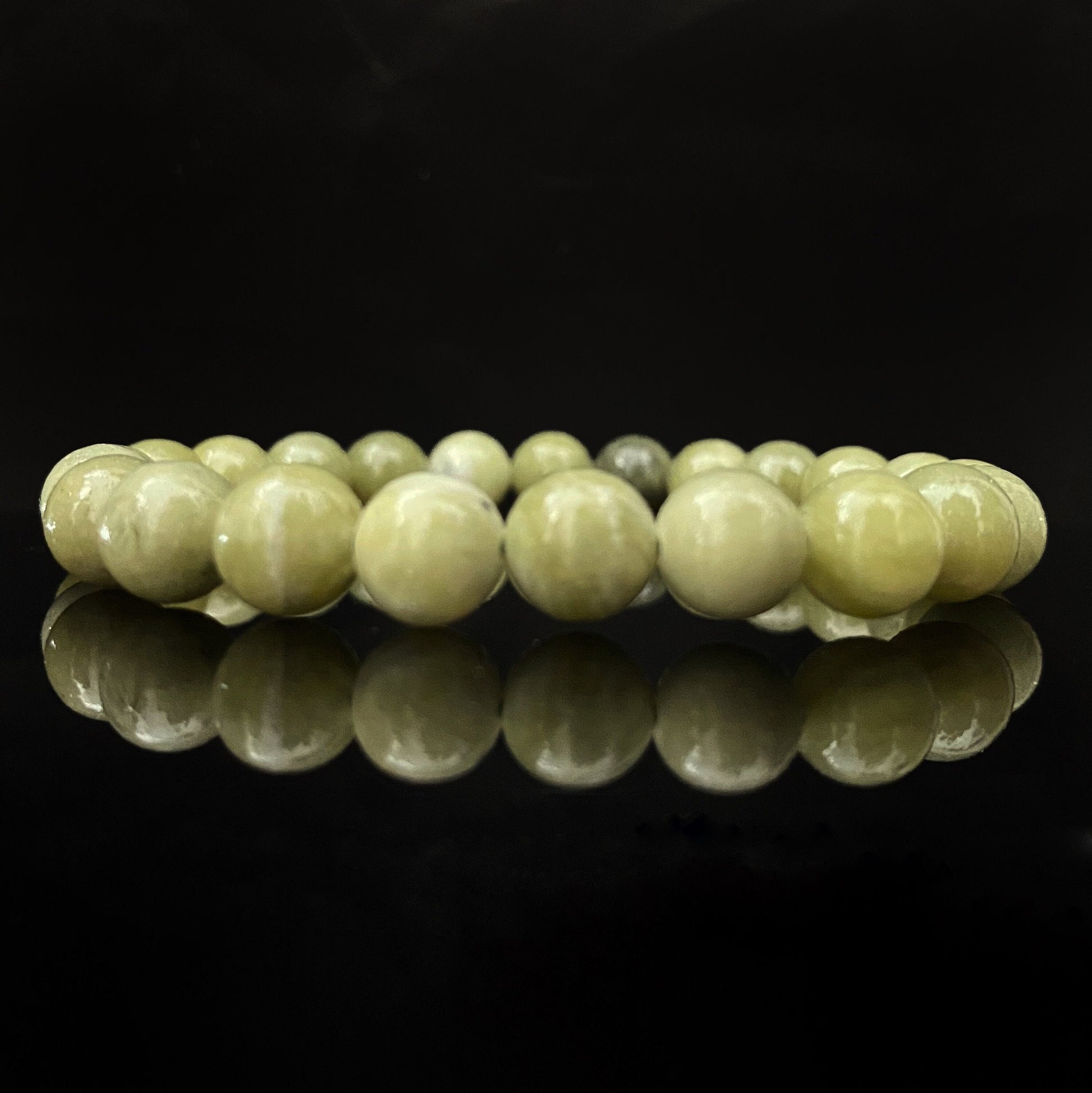 A polished 8mm Chinese Jade Beaded Bracelet forms a closed loop against a solid black background. The smooth, round jade polished beads are a pale green color, and the bracelet's reflection is clearly visible on the shiny surface below.
