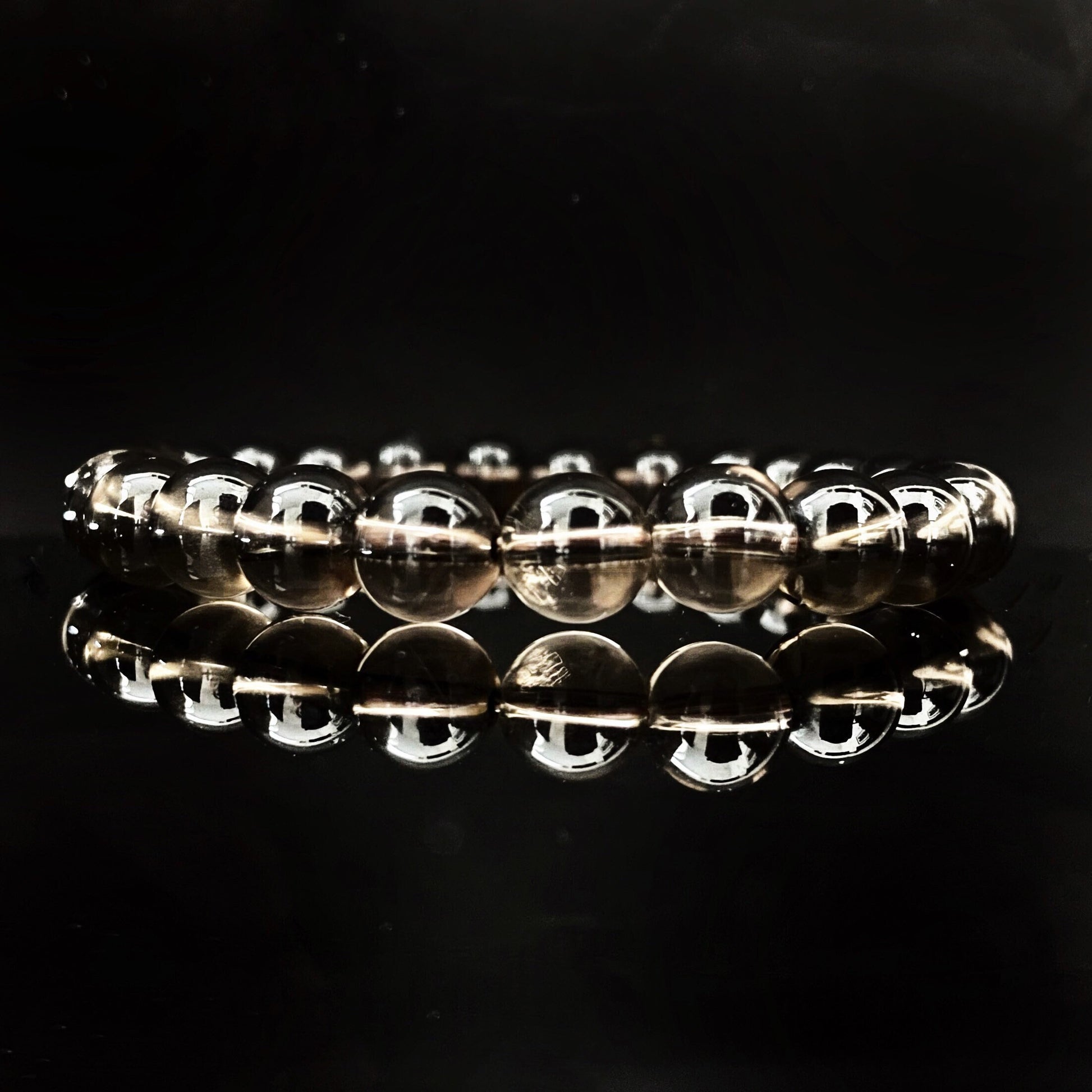 A close-up image shows several stacked 8mm Smoky Quartz Beaded Bracelets made of round, translucent beads. In the background, there are decorative elements, including small off-white and red dried flowers against a plain, light-colored surface. These grounding stone bracelets exude calming Usui Reiki energy.