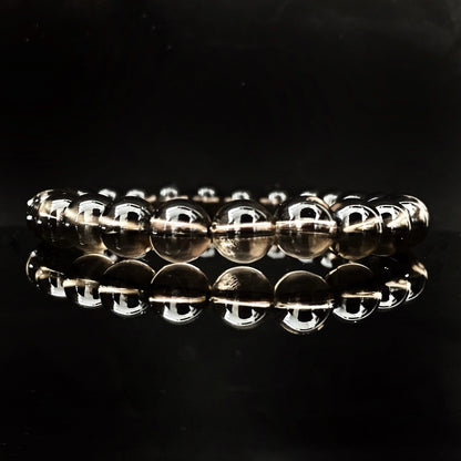 A close-up image shows several stacked 8mm Smoky Quartz Beaded Bracelets made of round, translucent beads. In the background, there are decorative elements, including small off-white and red dried flowers against a plain, light-colored surface. These grounding stone bracelets exude calming Usui Reiki energy.