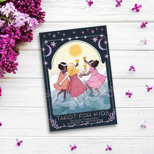 A children card deck titled "Tarot for Kids" is placed on a white wooden surface with scattered purple flowers and lilac blossoms around. The cover features three children in pink dresses dancing under a yellow sun, with a dark blue sky and celestial designs, perfect for enhancing decision-making skills.
