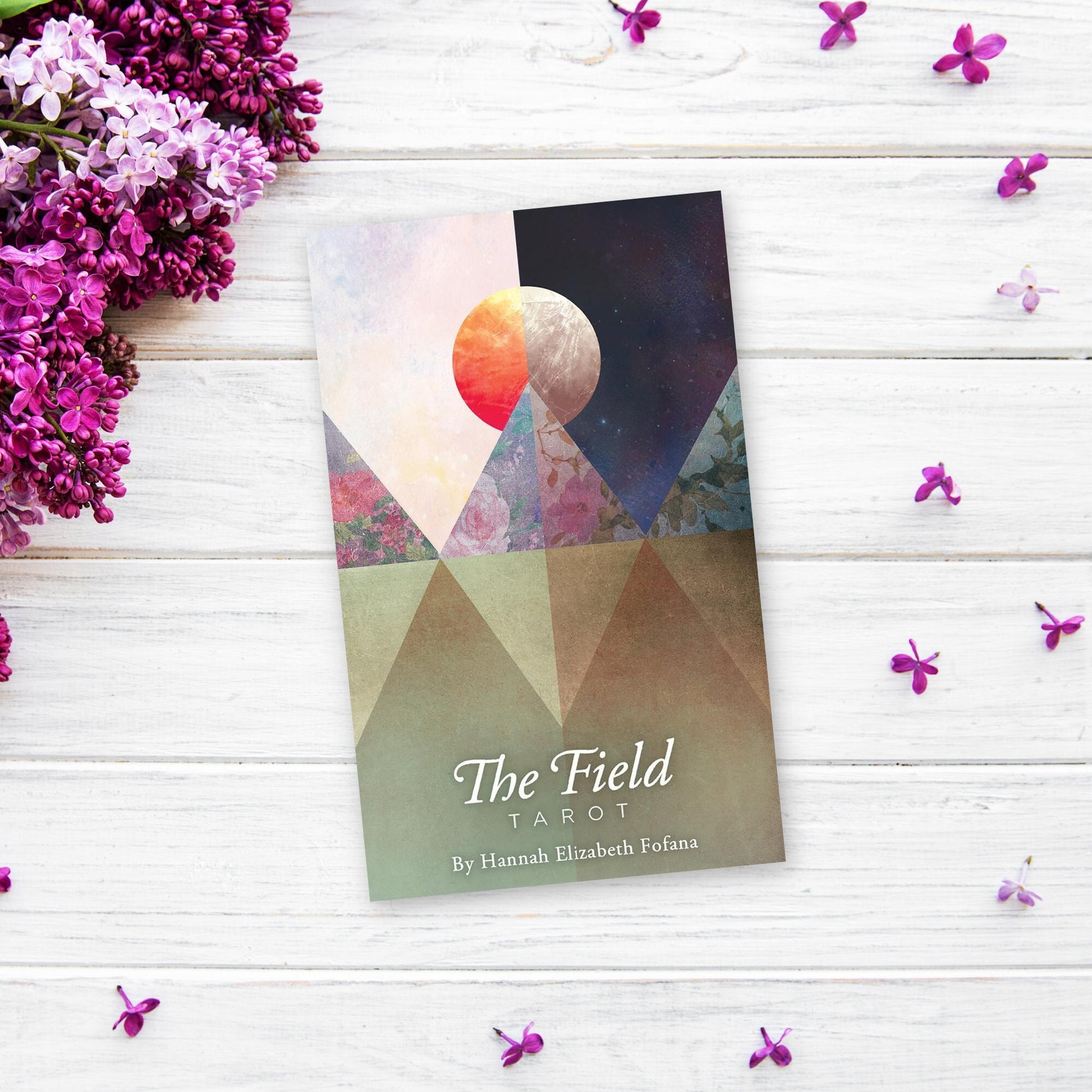 The image shows "The Field Tarot" by Hannah Elizabeth Fofana, placed on a white wooden surface. The cover features a stylized moon and mountains in earthy tones and vibrant colors. Scattered around the tarot deck are purple flowers and petals, evoking the essence of this intuitive tarot deck.