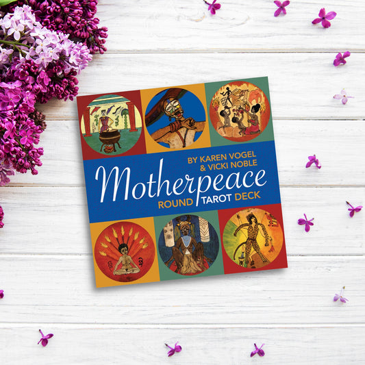 A box of Motherpeace Round Tarot Deck lays on a white wooden surface, surrounded by small pink flowers and sprigs of purple blossoms. The box features round cards with various illustrations, celebrating the spirit of a feminist tarot deck.