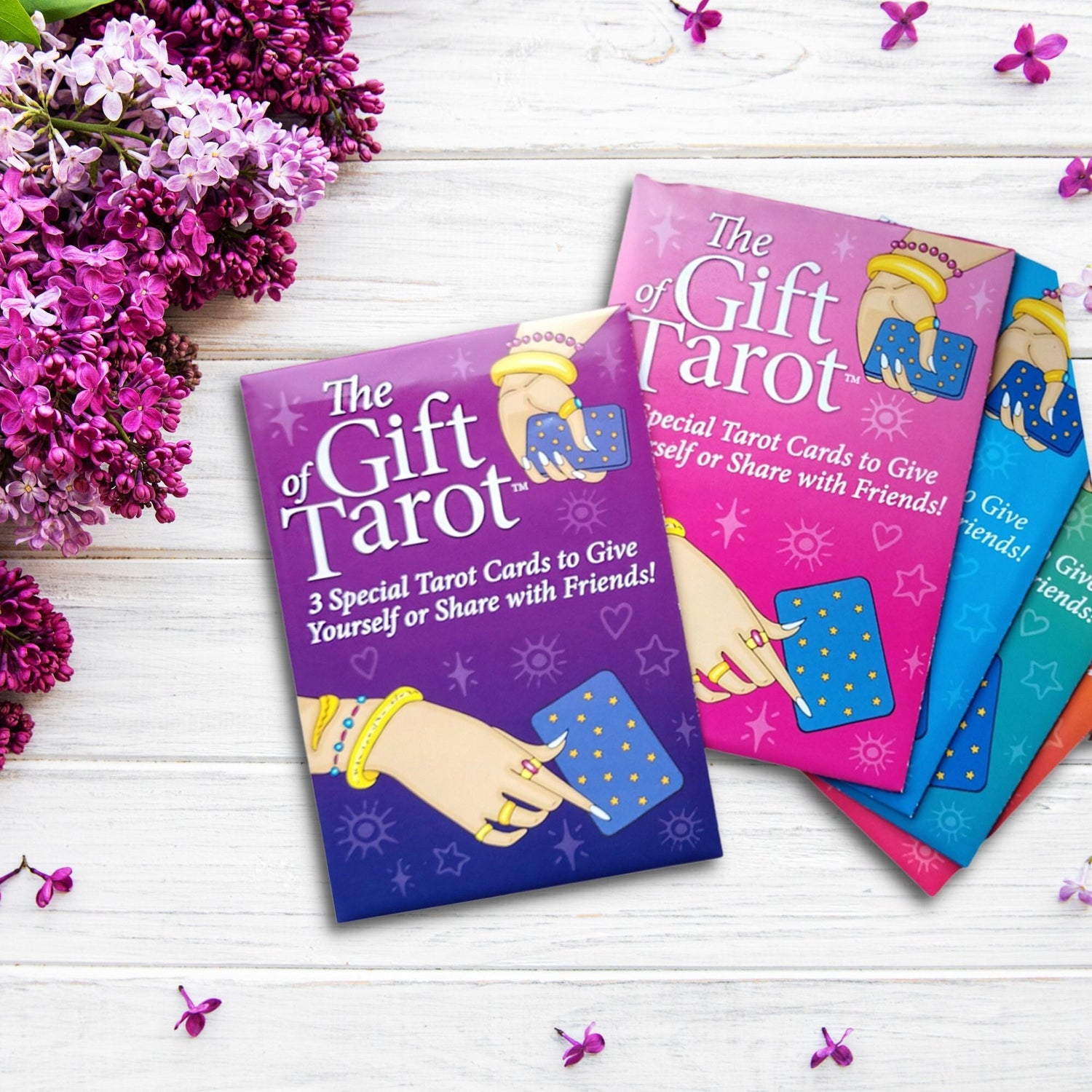 Three tarot card packages labeled "The Gift of Tarot (3 Card Tarot Reading Pack)," each featuring an illustration of a hand with jewelry holding a tarot card. The purple, pink, and green packages, reminiscent of the Radiant Rider-Waite style, rest on a white wooden surface adorned with purple lilac flowers on the left side.