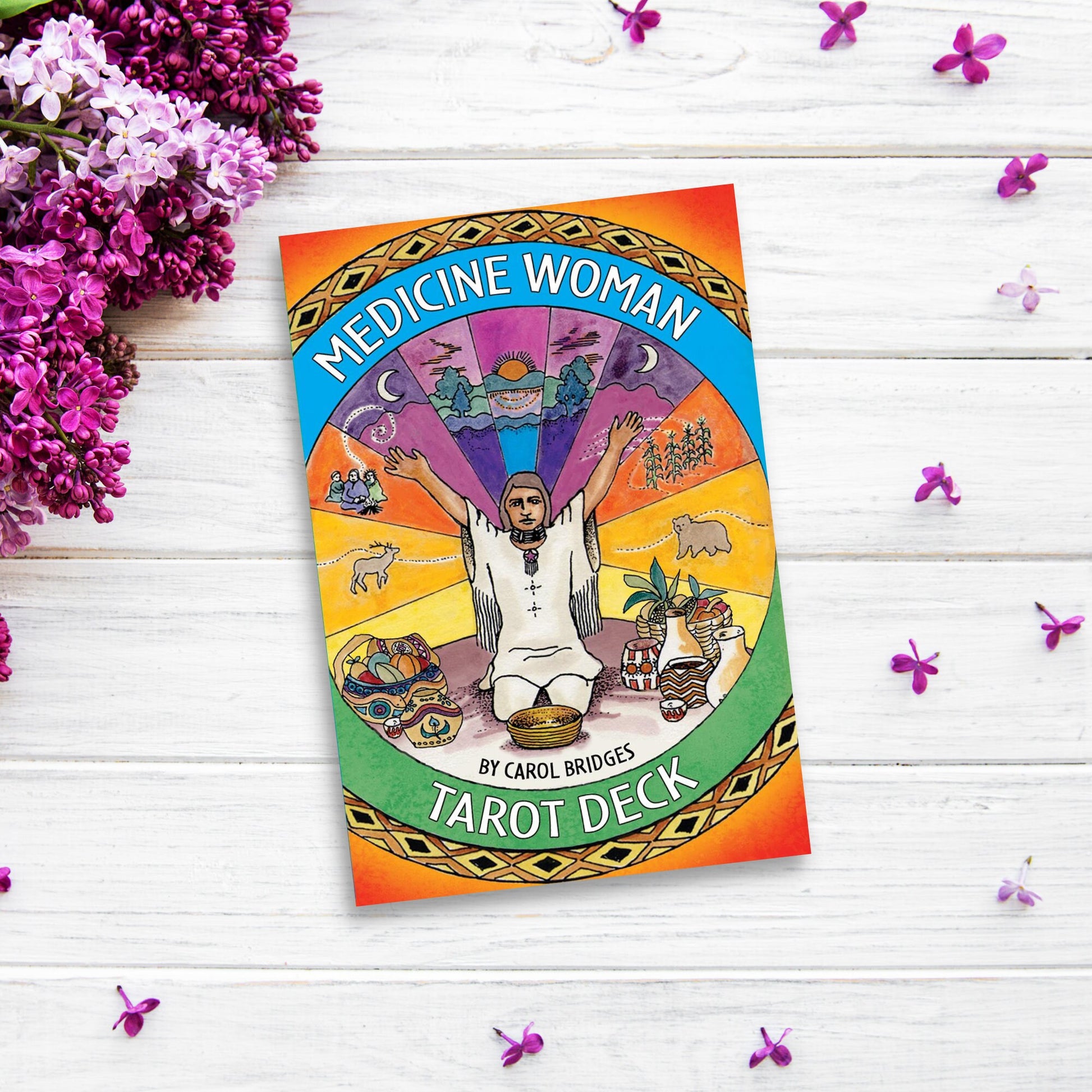The image shows a colorful "Medicine Woman Tarot" by Carol Bridges on a white wooden surface, surrounded by scattered purple flowers. This 78-card deck's cover features a woman with arms raised in front of a rainbow background and various symbolic images.