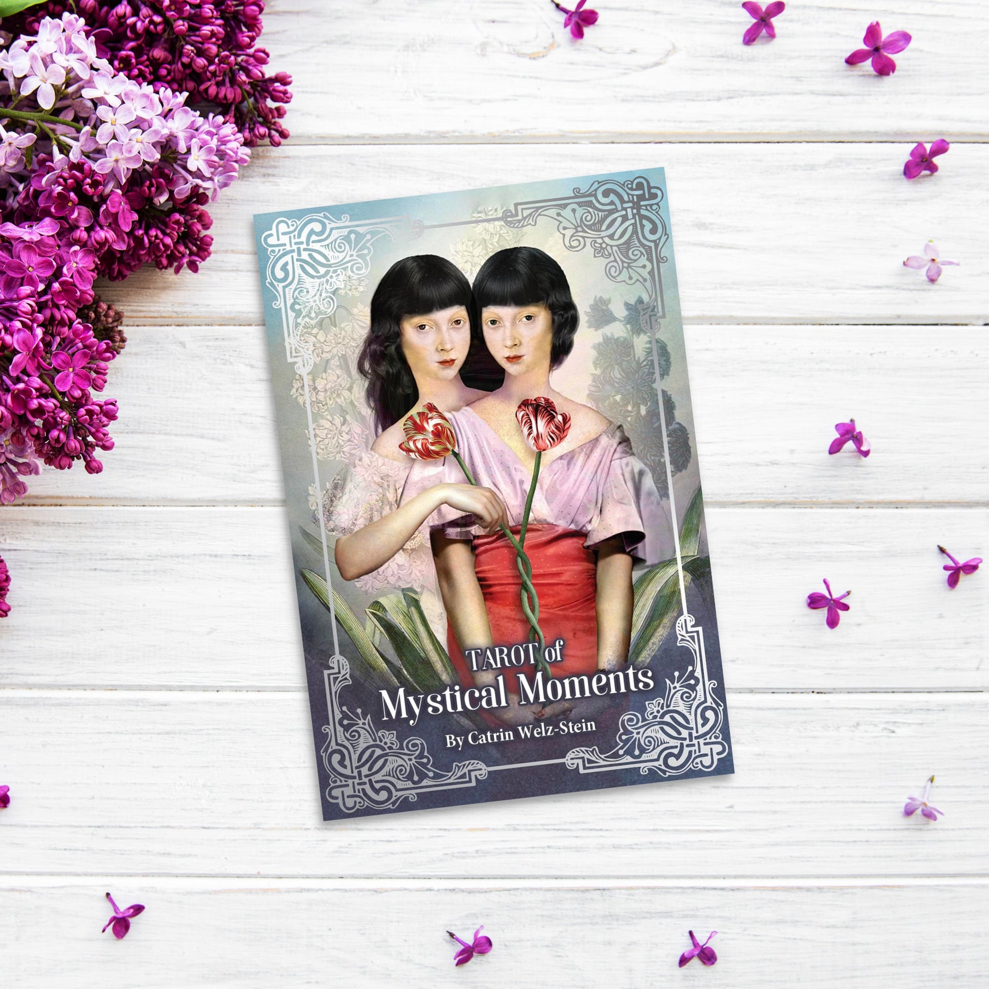 The image shows a "Tarot of Mystical Moments" deck by Catrin Welz-Stein on a white wooden surface. The cover depicts two women with blunt haircuts, one in a pink dress and the other in red, holding flowers. Purple and white flowers elegantly surround the "Tarot of Mystical Moments" tarot deck.
