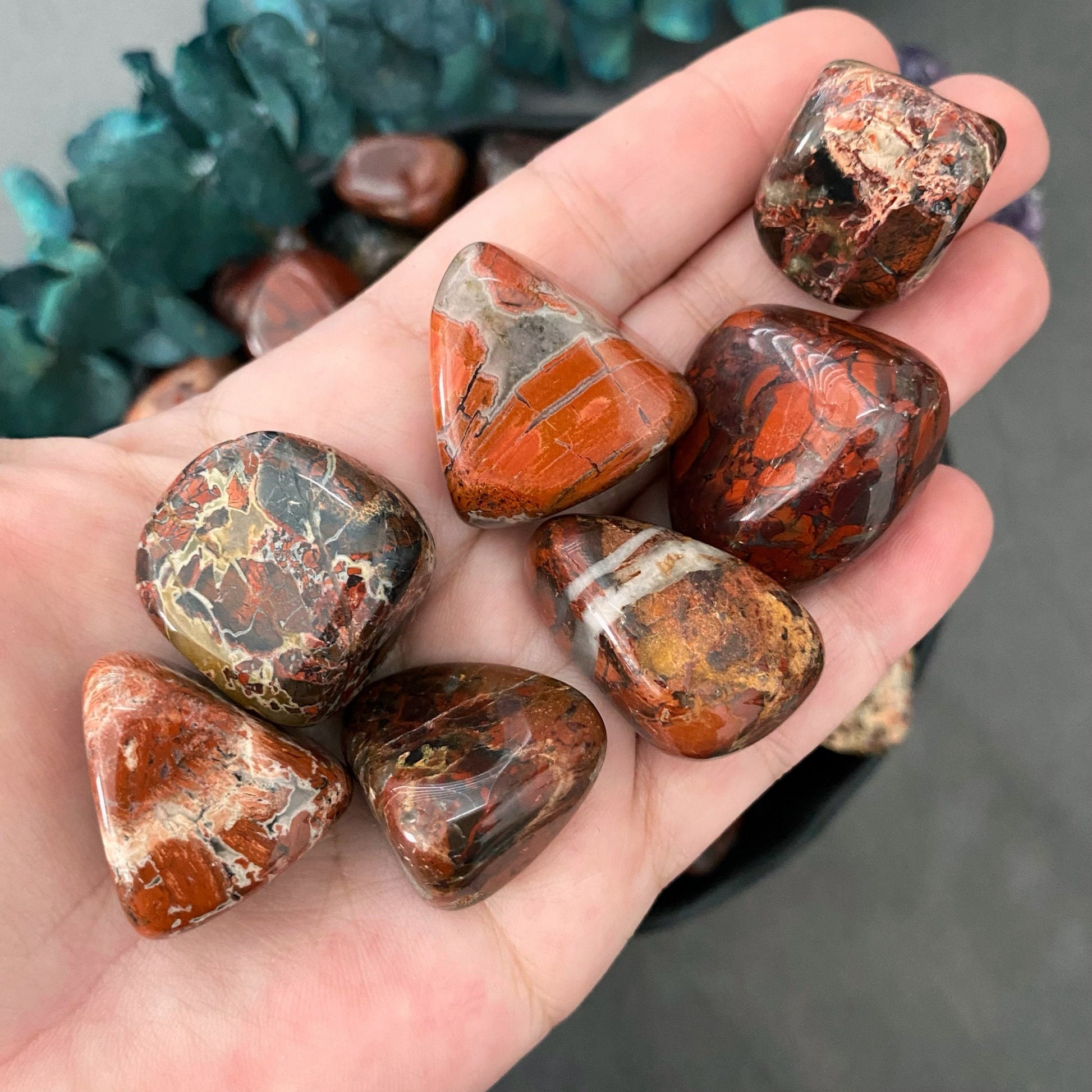 A black bowl filled with polished, multicolored jasper stones, predominantly in earthy tones such as reds, browns, and oranges, sits on a gray surface. The Brecciated Jasper Tumbled Stones are known for their grounding energy. To the left of the bowl, there is a small bunch of red, dried flowers.