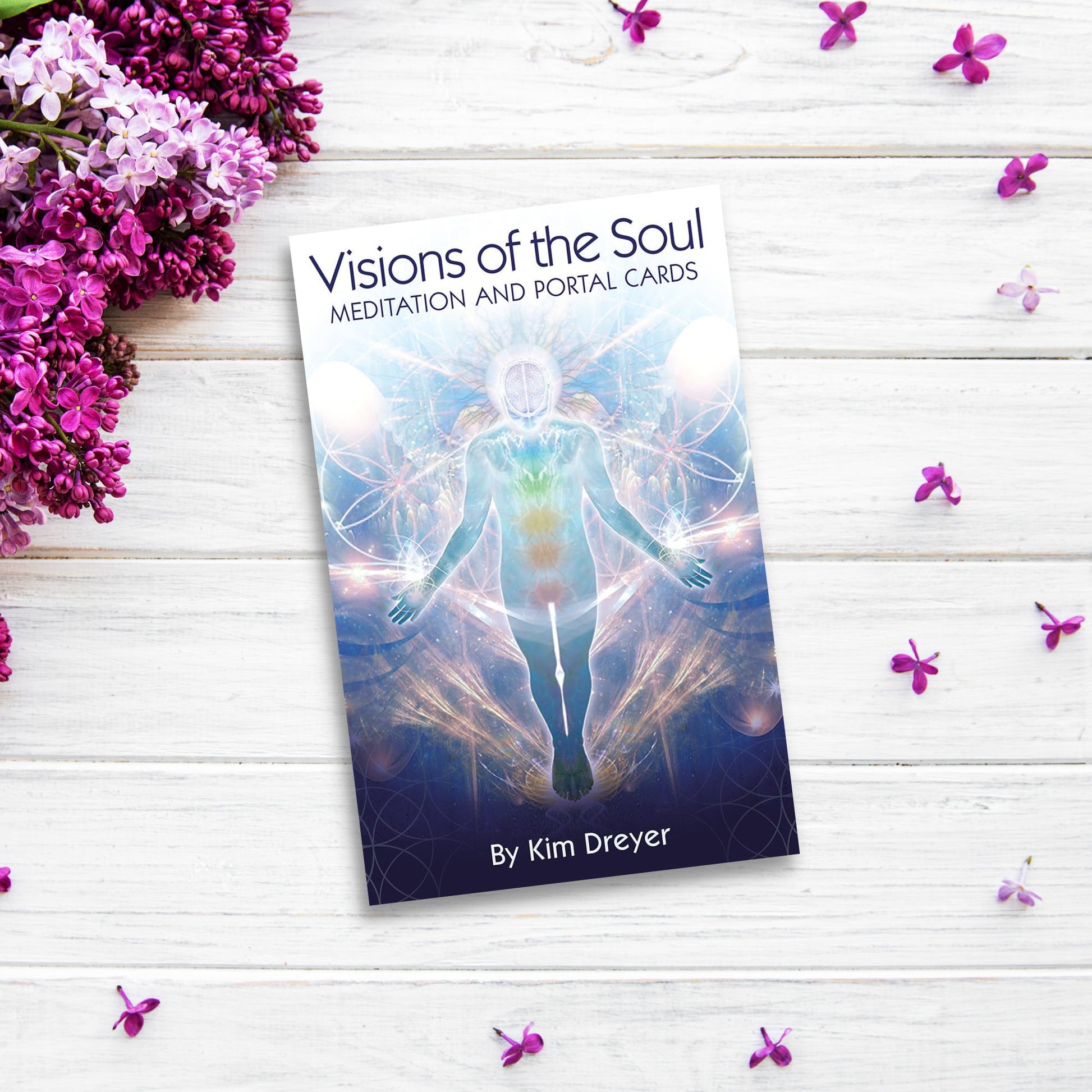 The product name is already correctly integrated into the sentence provided:

"A book titled "Visions of the Soul: Meditation and Portal Cards" by Kim Dreyer lies on a white wooden surface adorned with scattered purple flowers. The cover features an ethereal figure surrounded by colorful, mystical patterns—an exquisite oracle deck for spiritual seekers. Purple flowers are placed on the left.